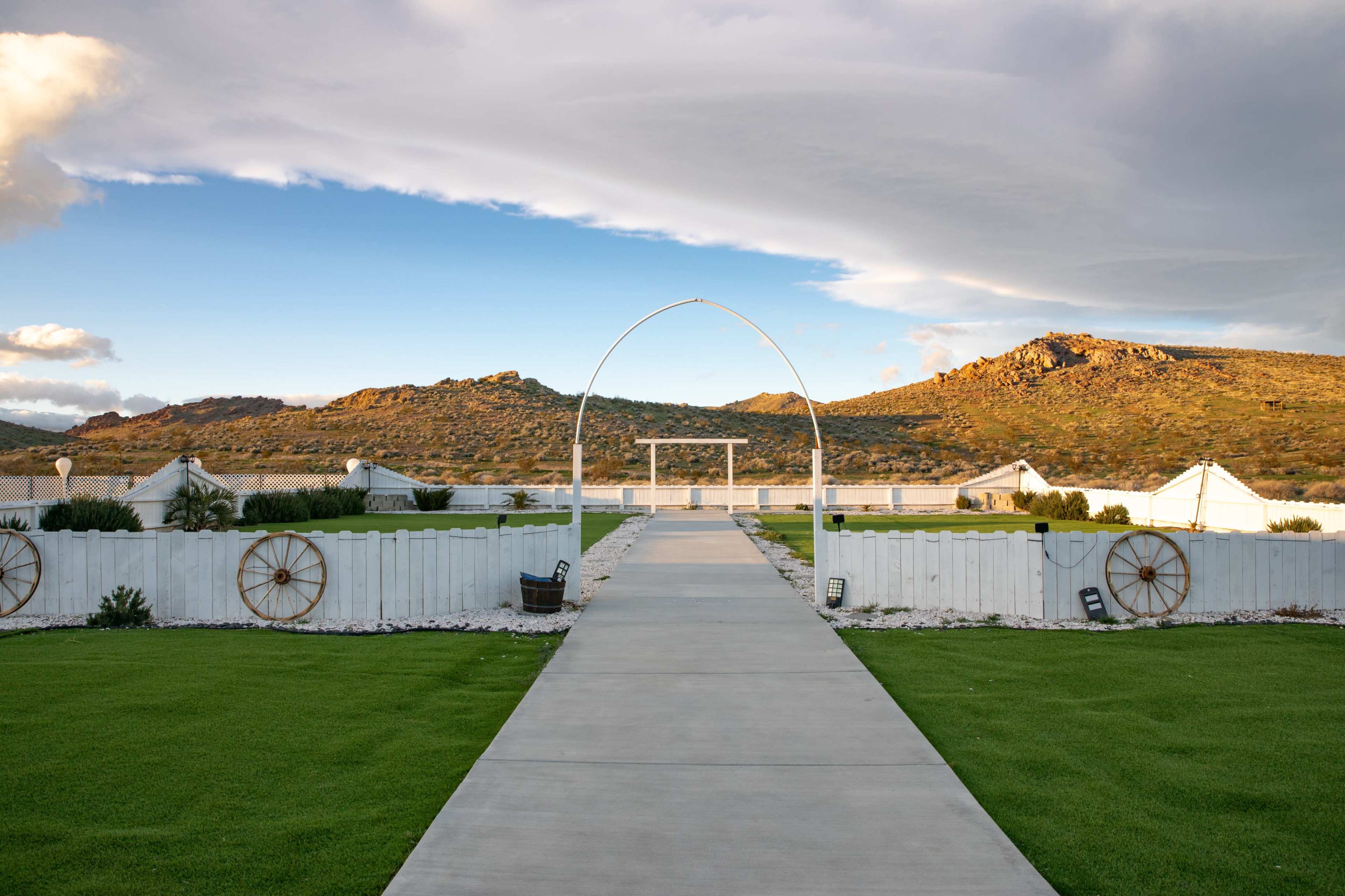The image shows a wide pathway leading through a landscaped area with a white arch and hills in the background under a partly cloudy sky.
