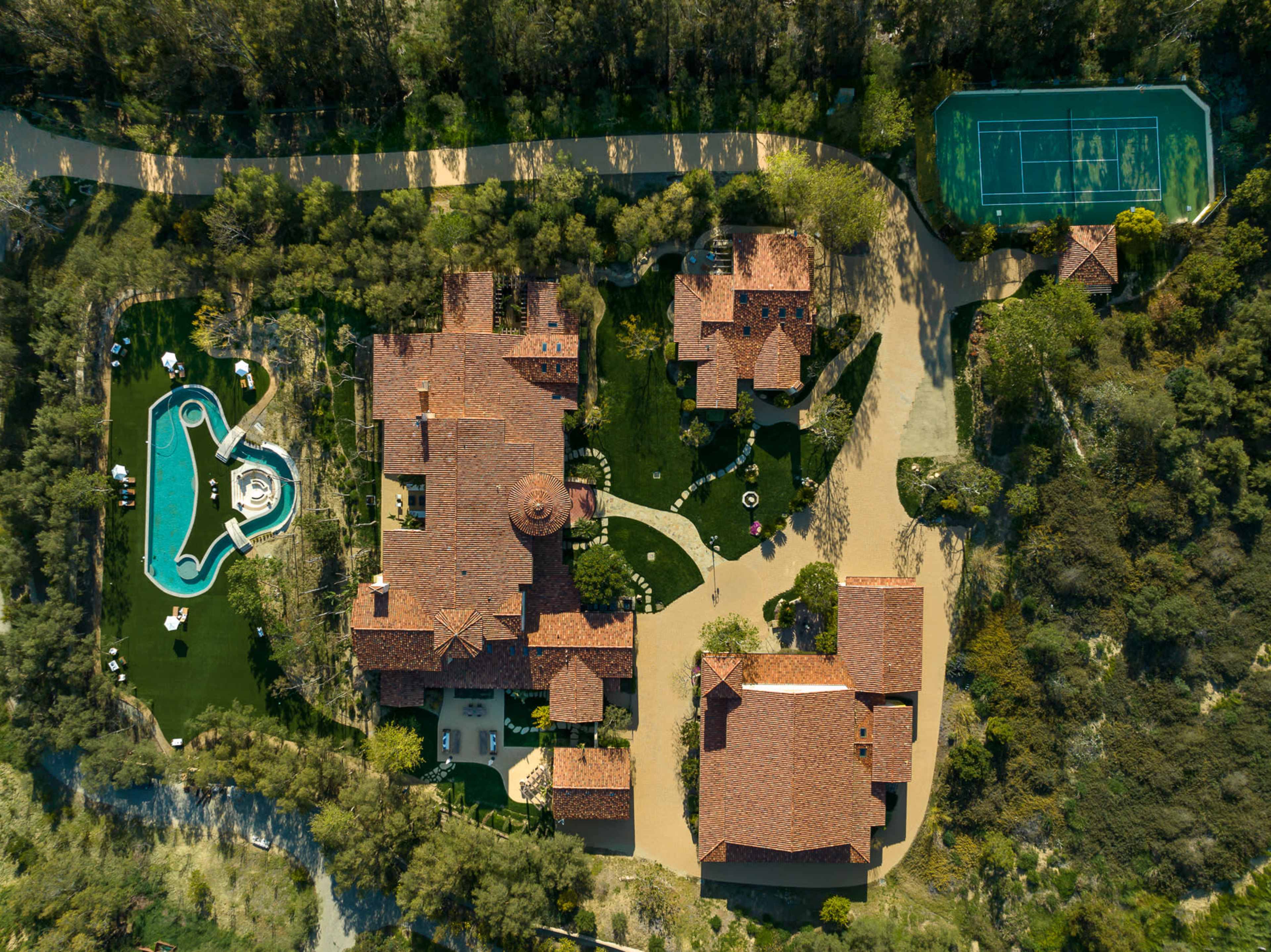 An aerial view of a large estate featuring a main house, guest houses, a swimming pool, and a tennis court set among trees.