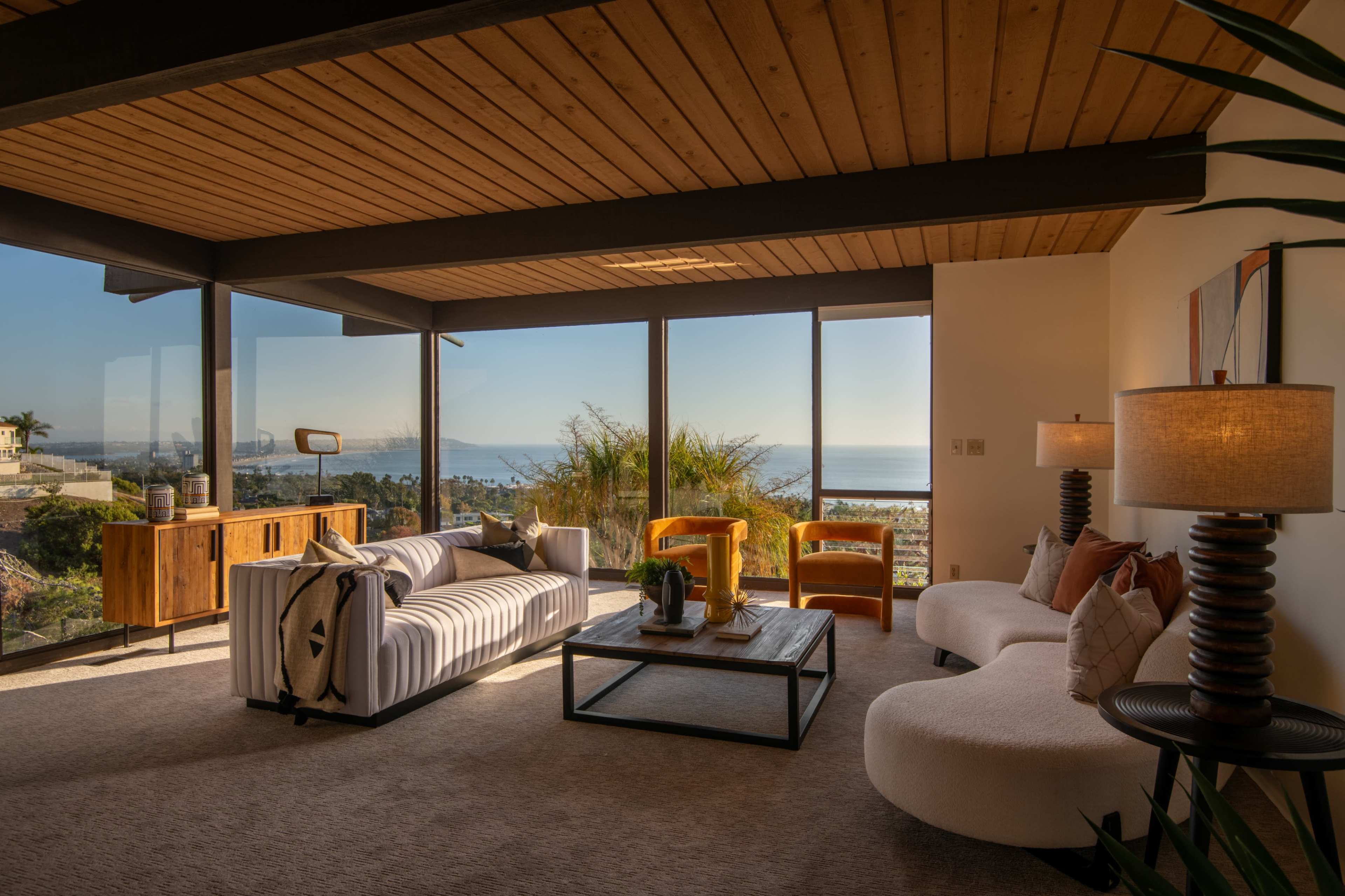 A modern living room features a large panoramic window with a view of the ocean, furnished with a white sofa, two curved chairs, a coffee table, and a wooden sideboard.