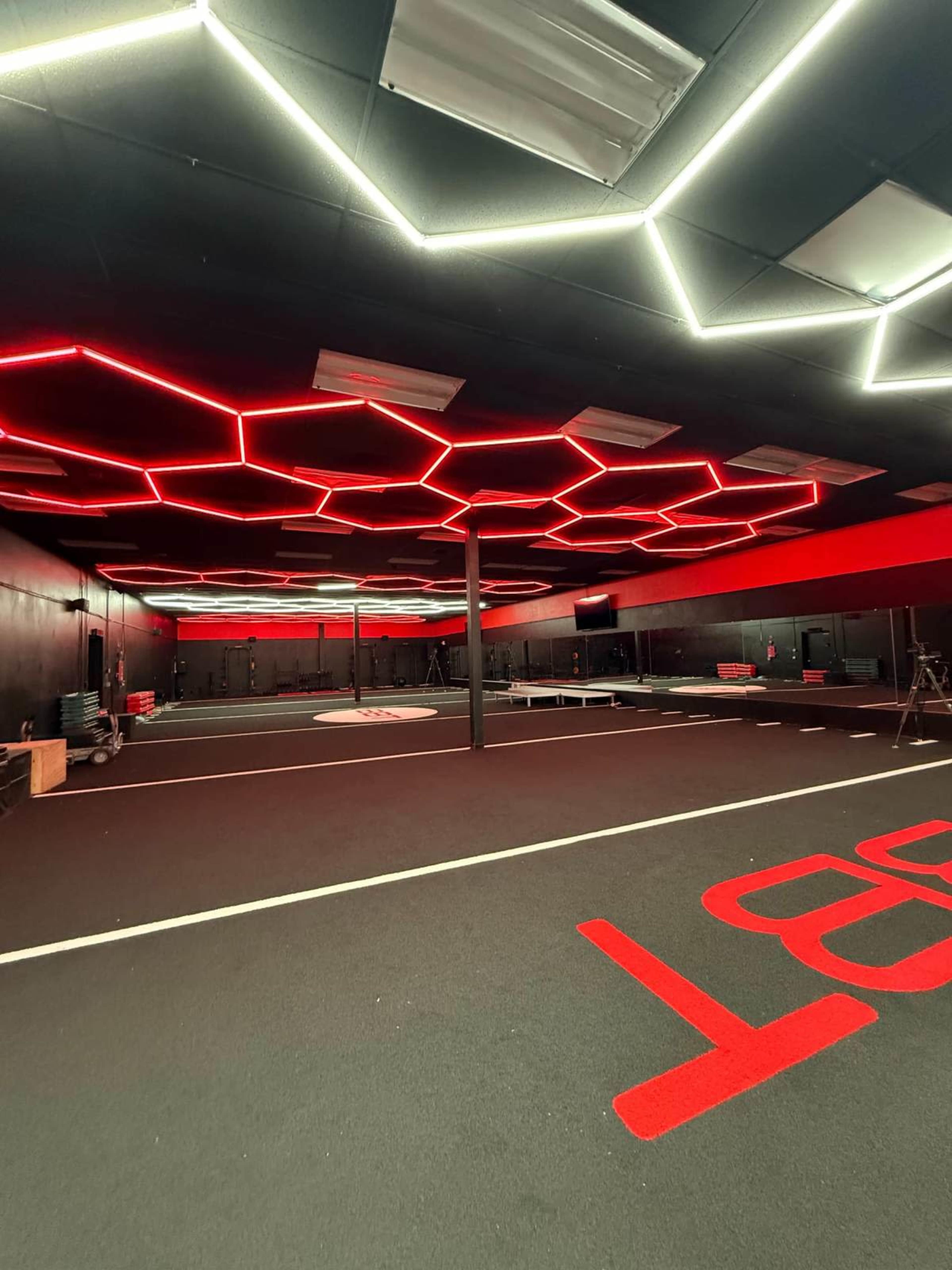 The image shows a modern gym interior with a black floor, hexagonal red lighting on the ceiling, and minimal equipment arranged in an open space.
