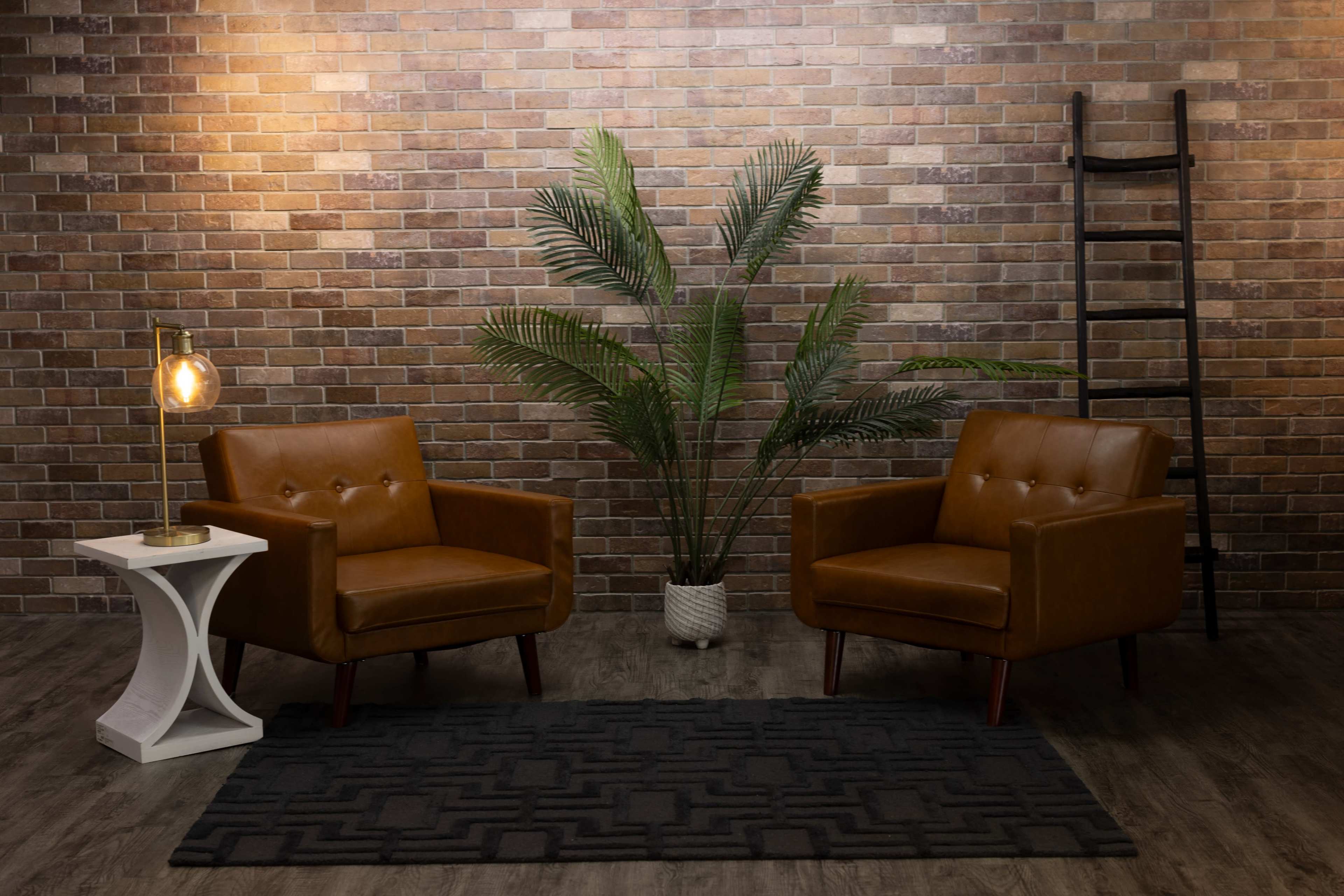 Two brown leather armchairs positioned facing each other in a room with a brick wall, accompanied by a palm plant, a side table with a lamp, and a decorative ladder.