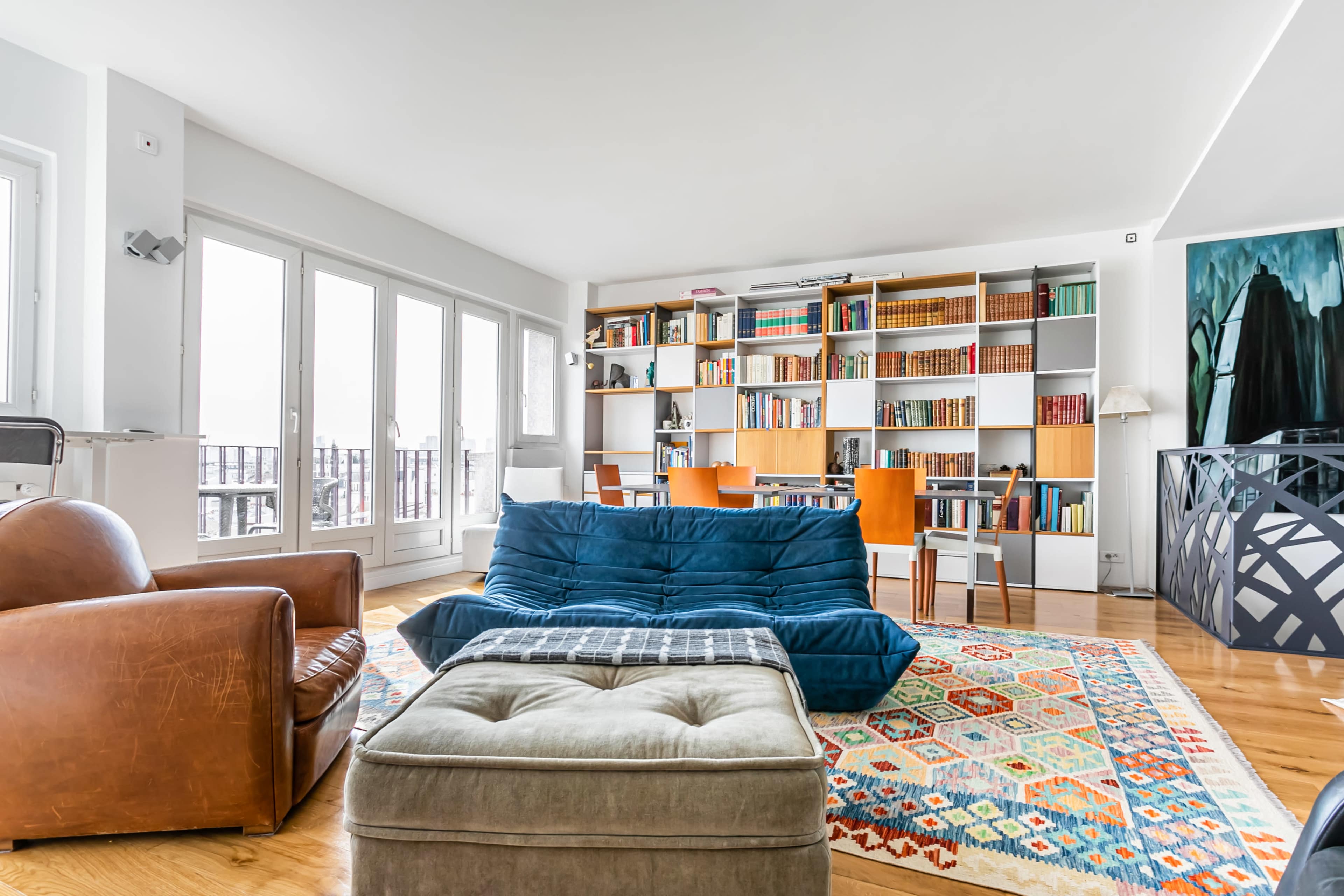 A contemporary living room with a blue sofa, a brown armchair, and a bookshelf filled with books in front of large windows.