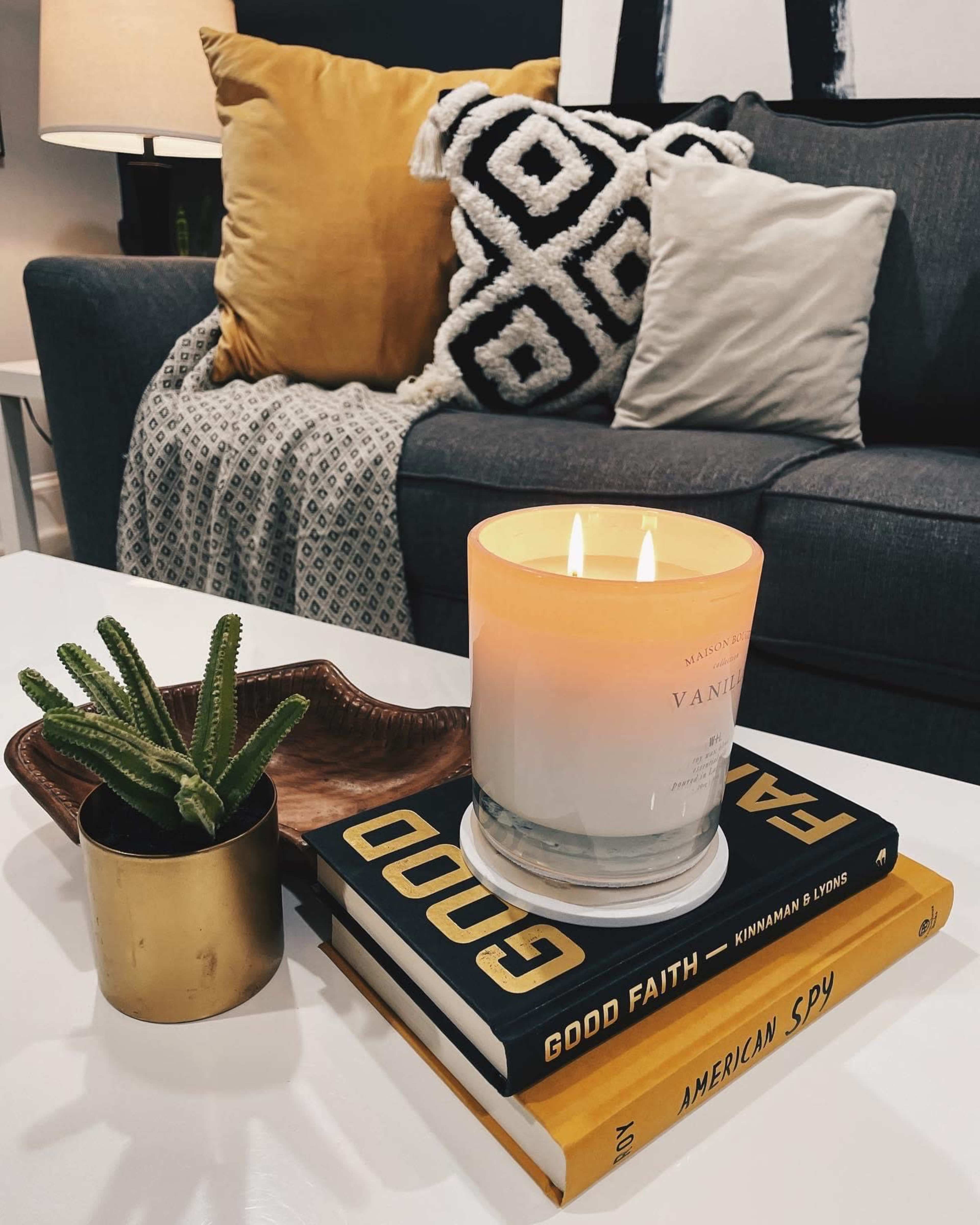 A lit candle sits atop a stack of books on a coffee table, accompanied by a small potted plant and a decorative tray, with a gray sofa and patterned cushions in the background.