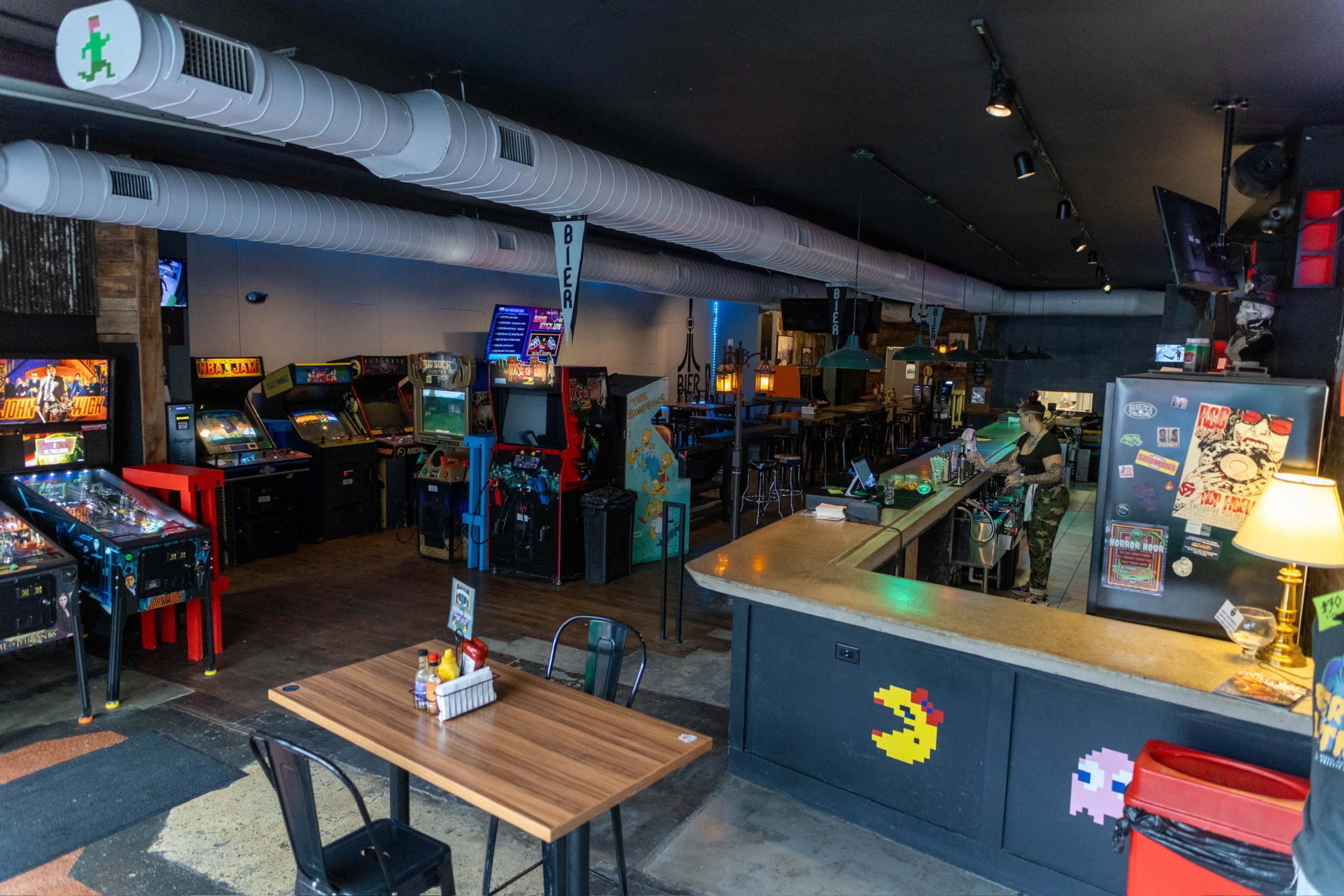 The image shows a retro arcade bar featuring various classic video game machines and a bar area with seating, illuminated by dim lighting.