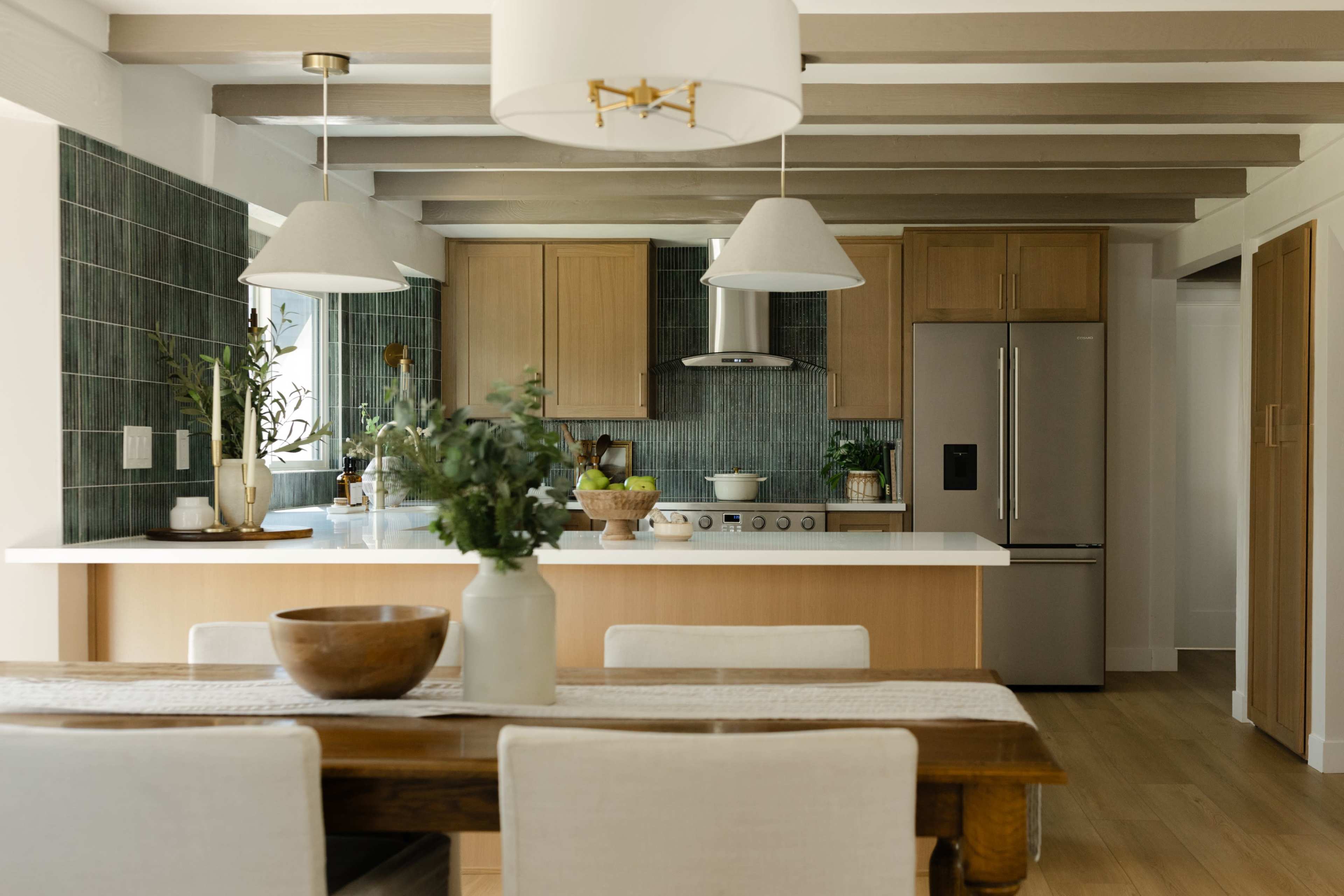 The image shows a modern kitchen with wooden cabinetry, a stainless steel refrigerator, and an adjacent dining area featuring a wooden table and white chairs.