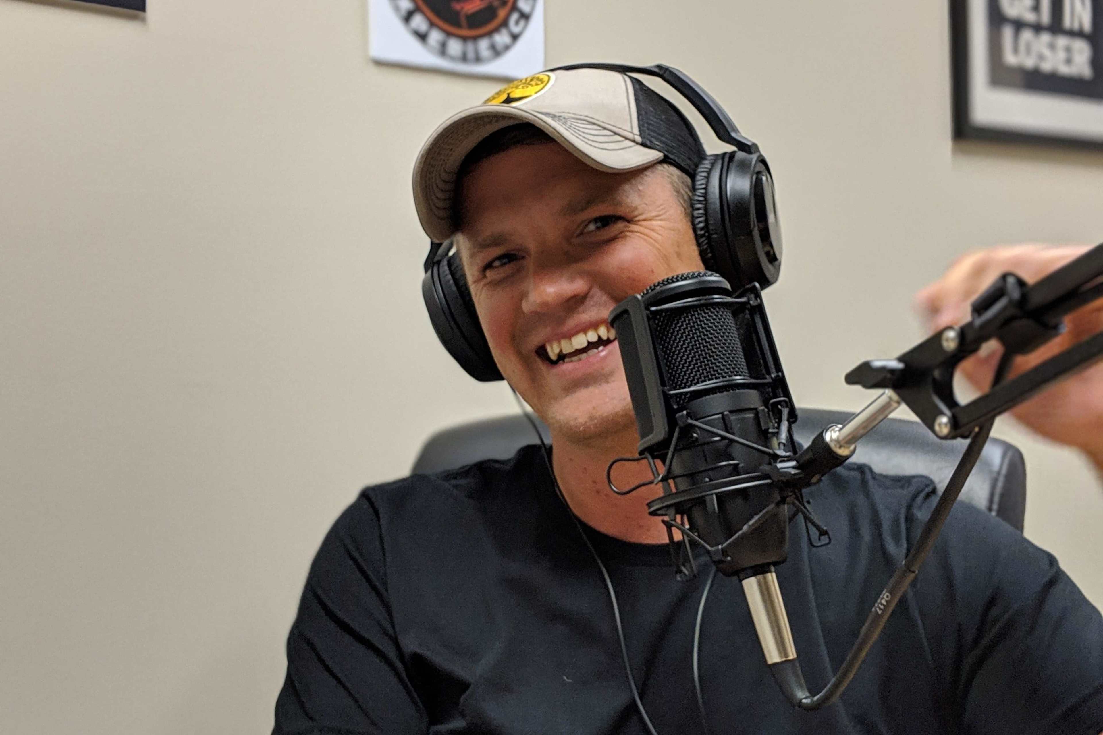 A person wearing a cap and headphones smiles while sitting in front of a microphone in a podcast studio.