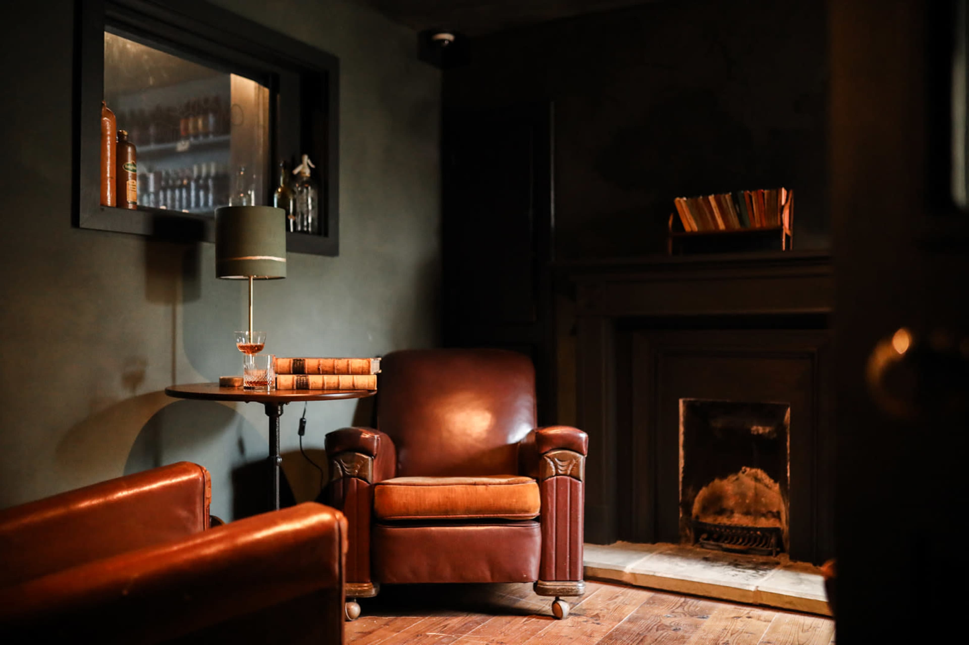 The image shows a dimly lit room featuring a leather armchair beside a small table with a lamp, a fireplace, and a shelf of books in the background.