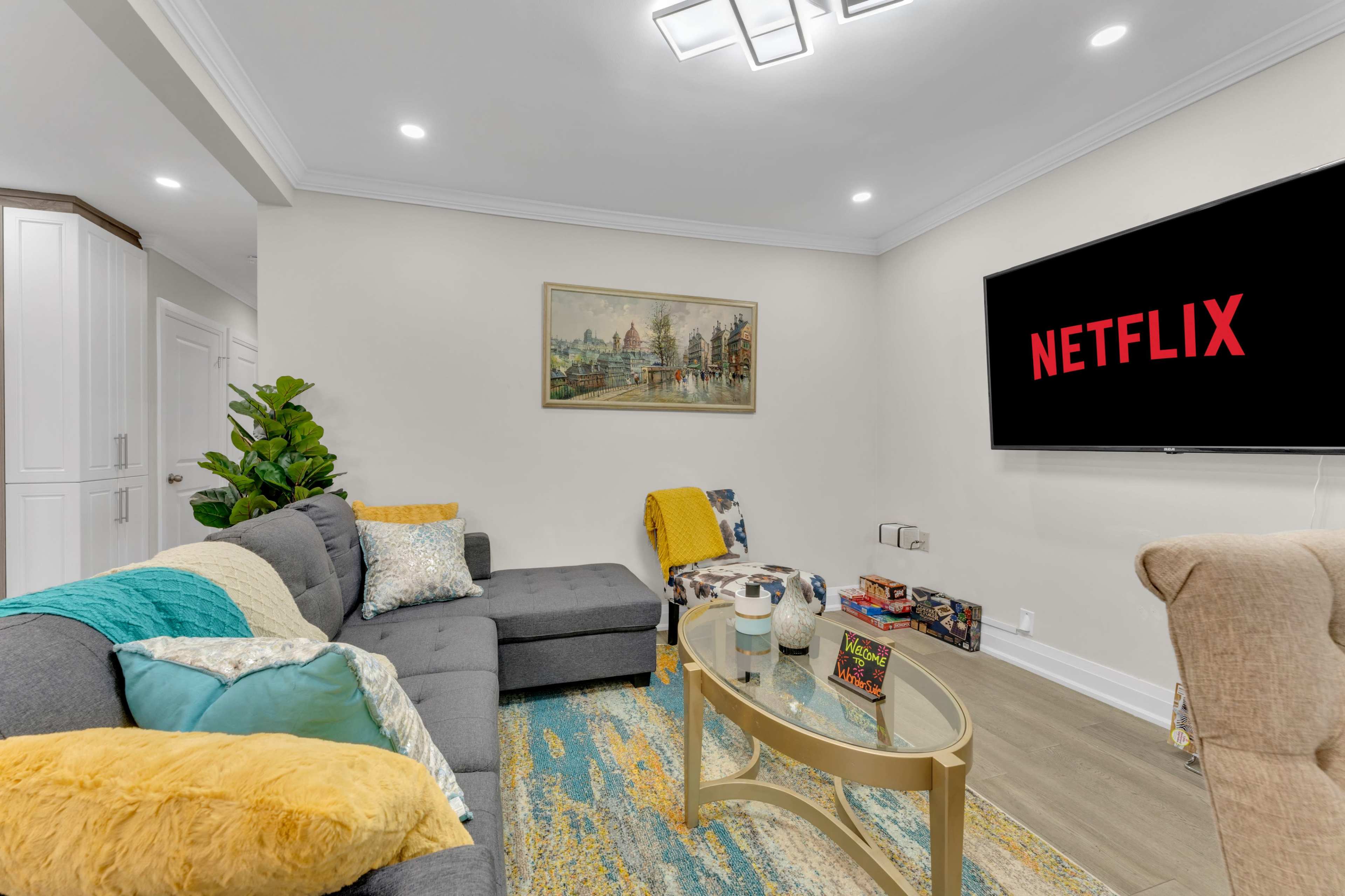 A modern living room features a gray sectional sofa, a glass coffee table, and a large television displaying the Netflix logo.