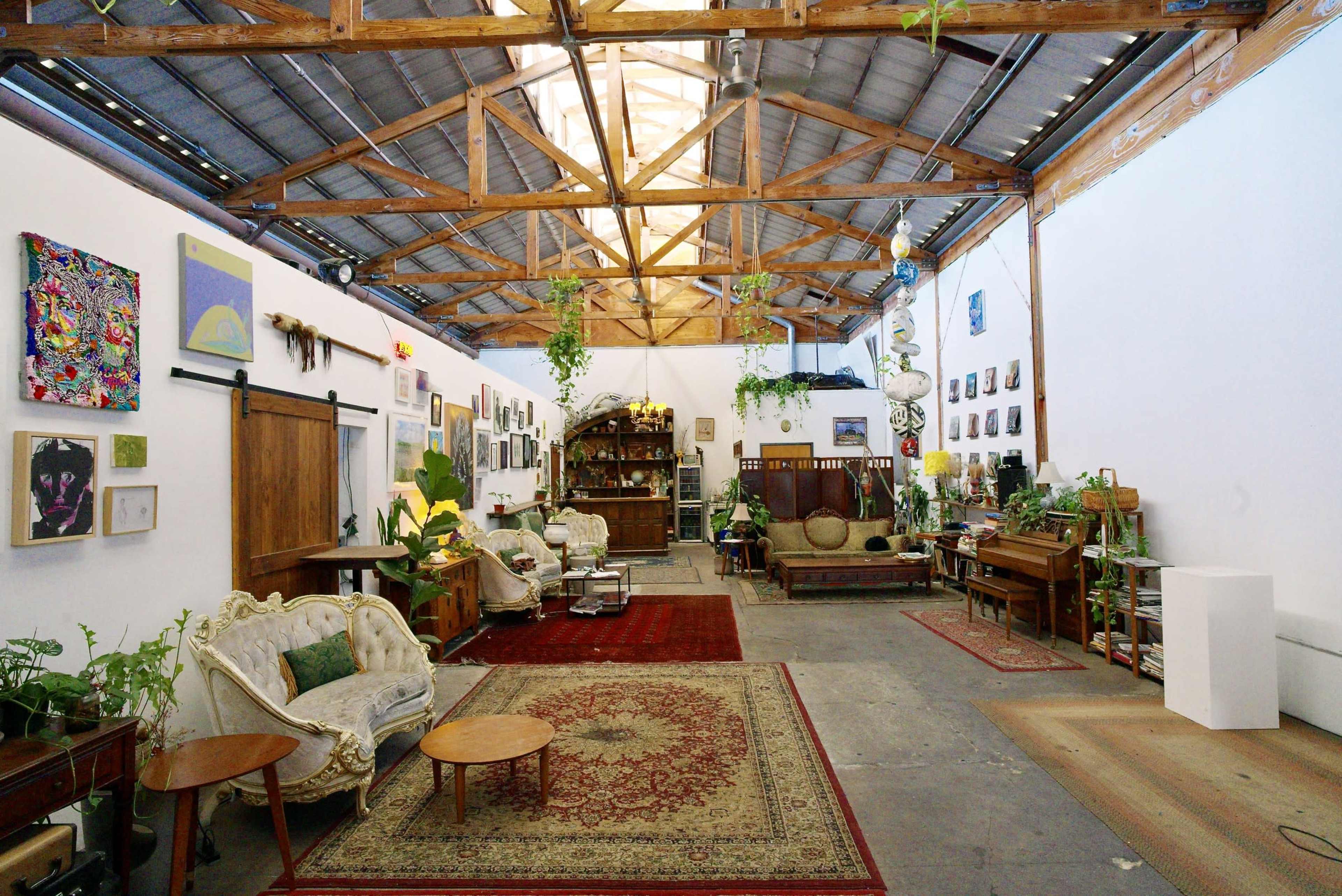 The image depicts a spacious loft-like interior with exposed wooden beams, eclectic furniture, and various plants, featuring artwork displayed on the walls.