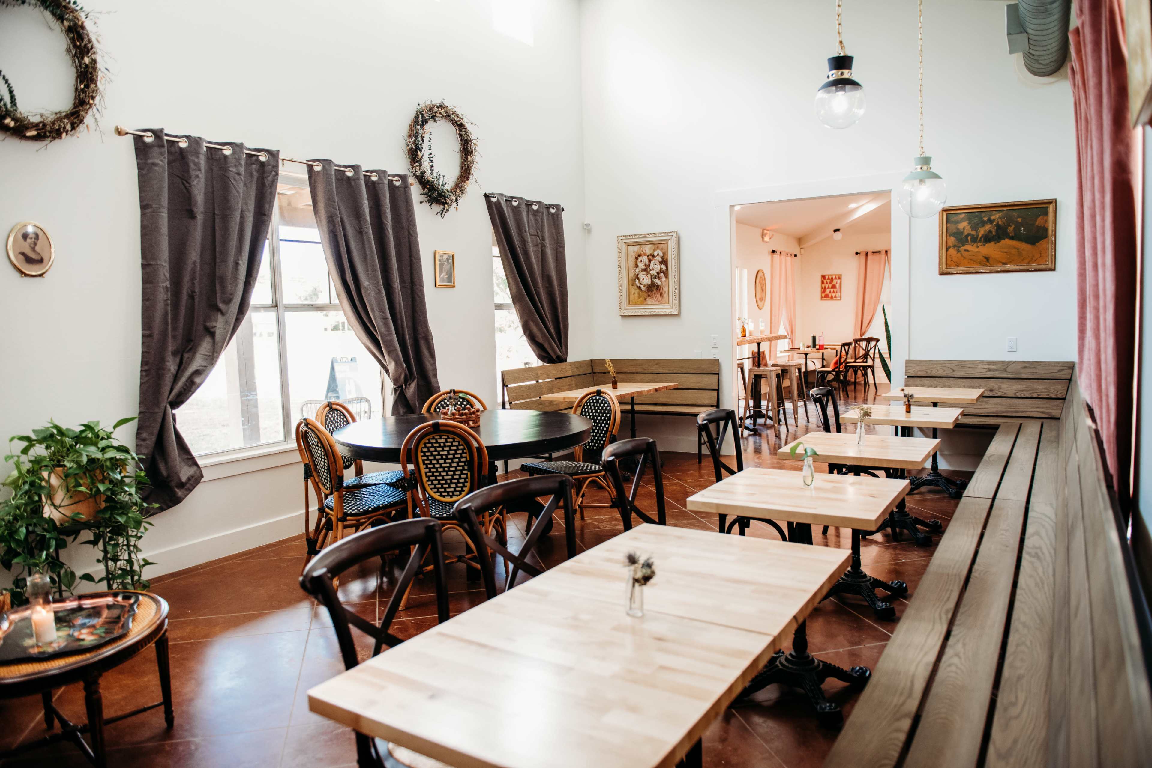 A bright café interior with wooden tables, chairs, and dark curtains, featuring a cozy seating arrangement and artwork on the walls.