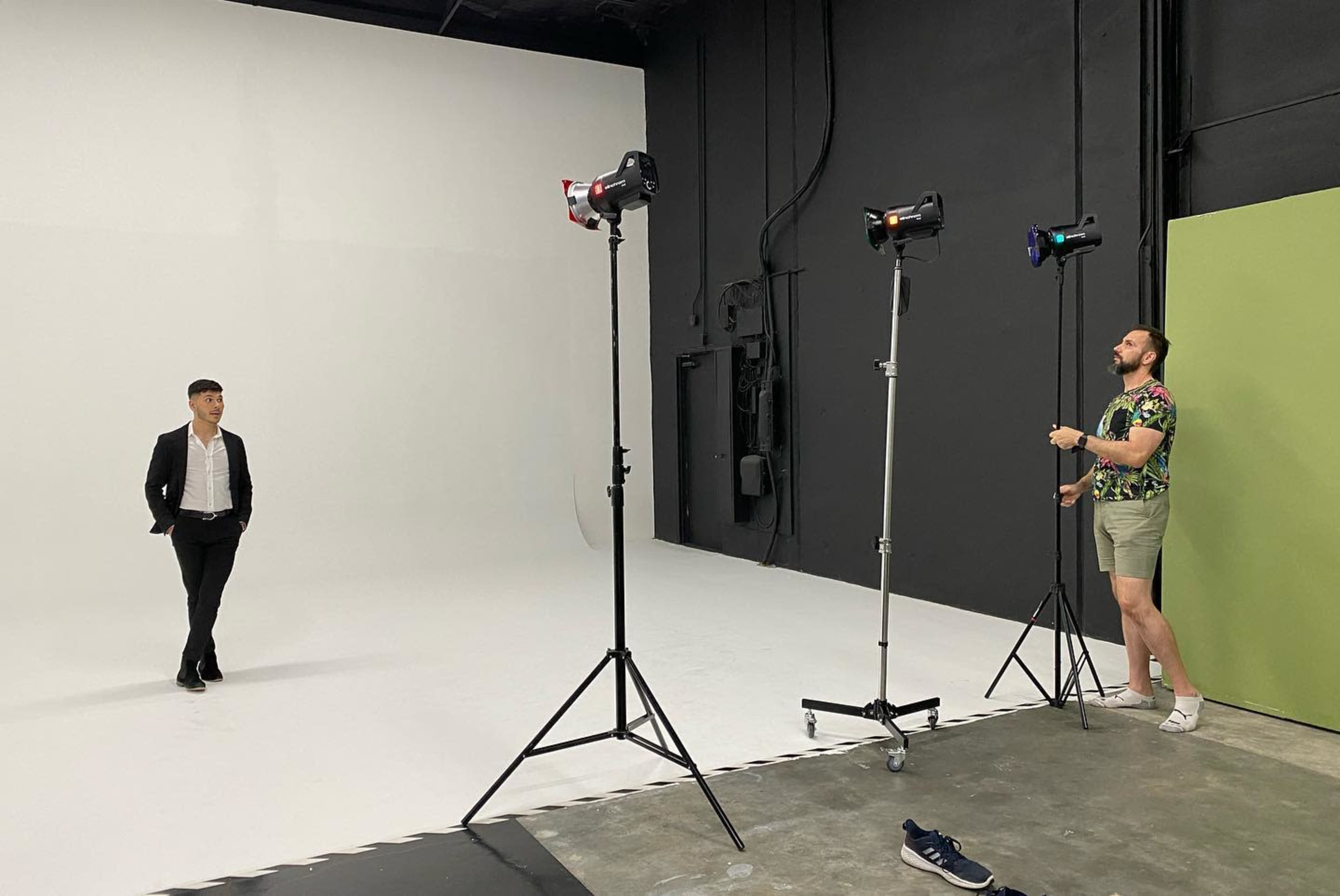 A man in a black suit stands in the center of a photography studio while another man adjusts lighting equipment nearby.