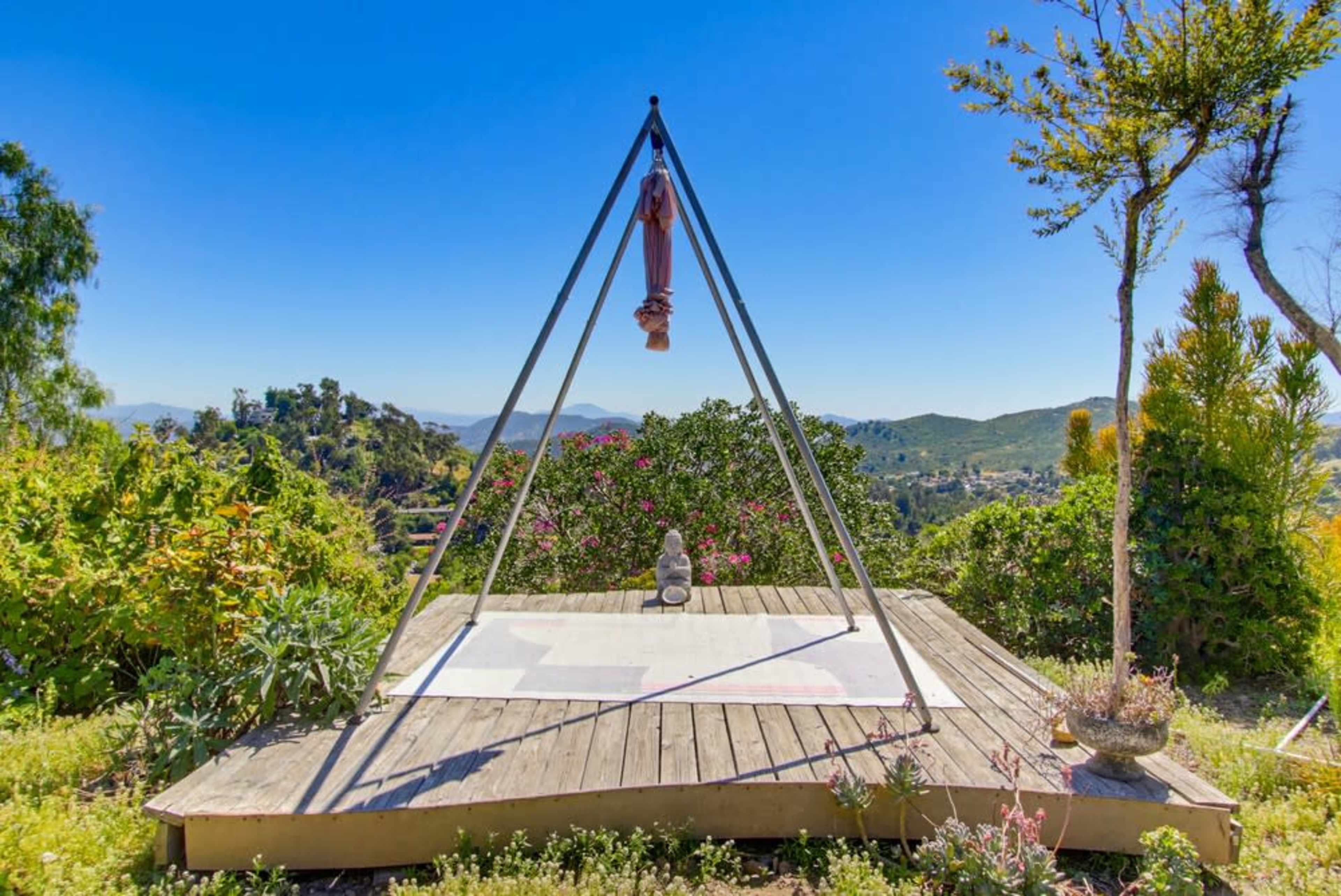 The image shows a triangular structure on a wooden platform surrounded by greenery and hills in the background.
