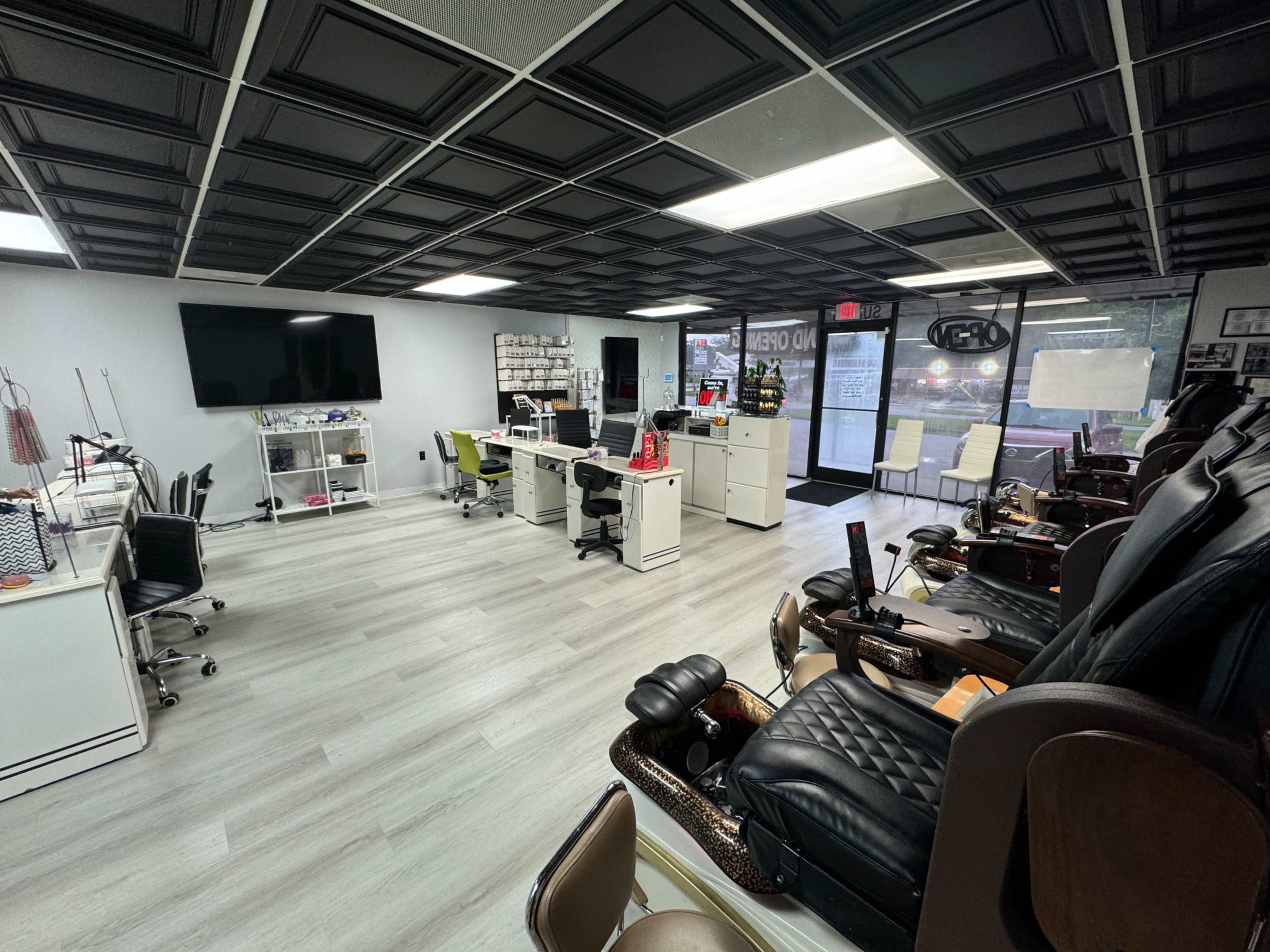 The image shows a spacious nail salon with several manicure stations, pedicure chairs, and a large window displaying a view of the street outside.