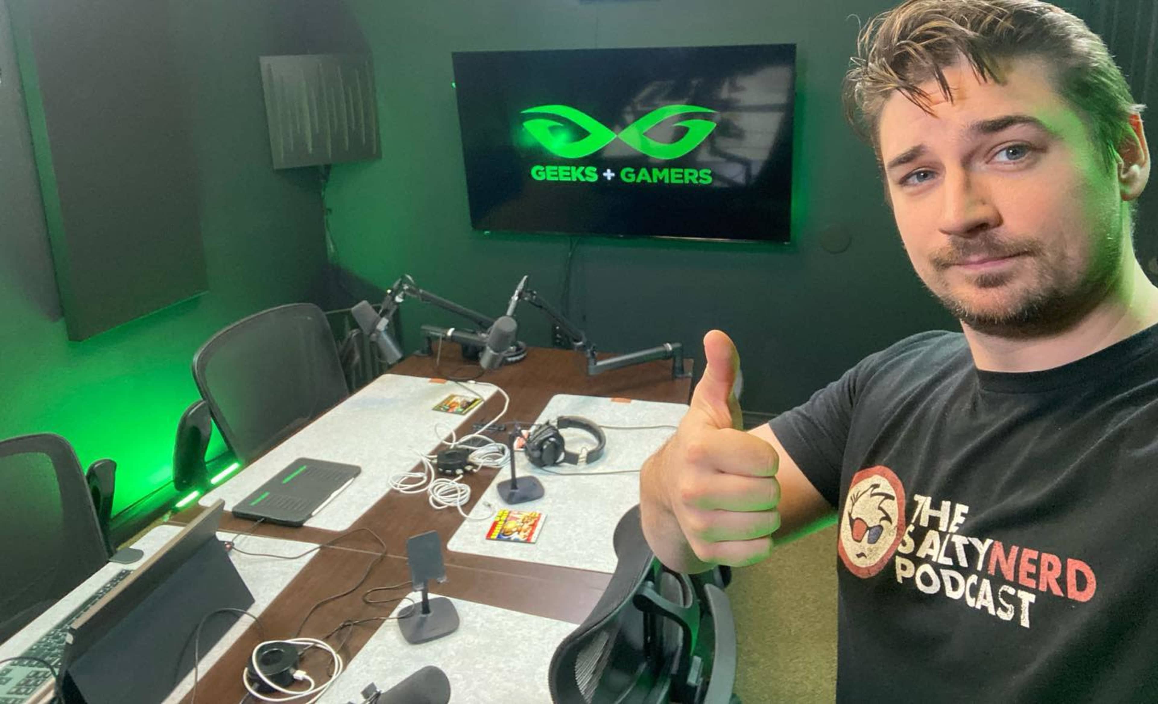 A man poses for a selfie in a podcast studio with green lighting and a large screen displaying the "Geeks + Gamers" logo.