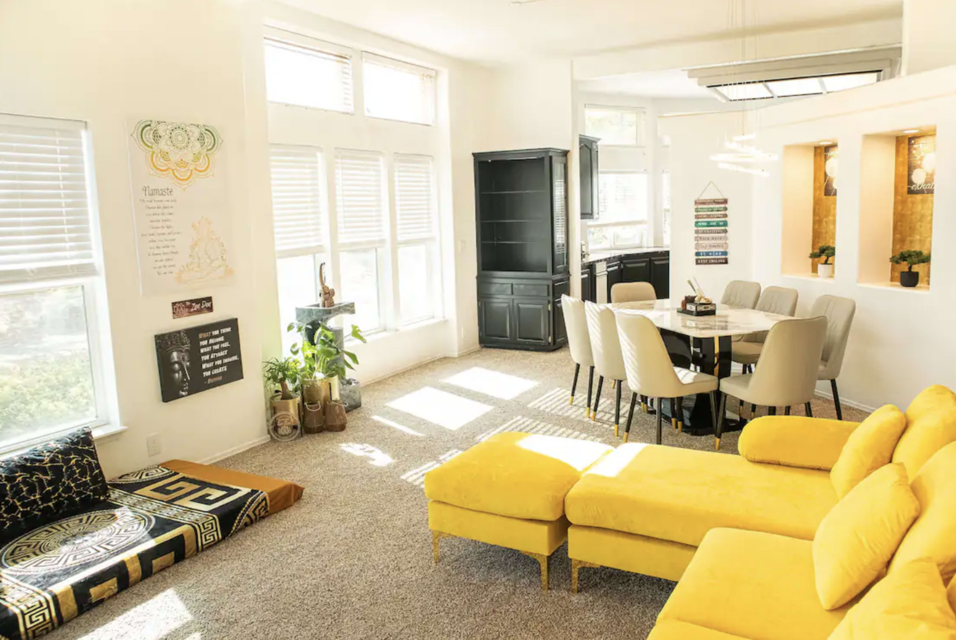 A bright living area features a yellow sectional sofa, a round dining table with gray chairs, and large windows allowing natural light to fill the space.