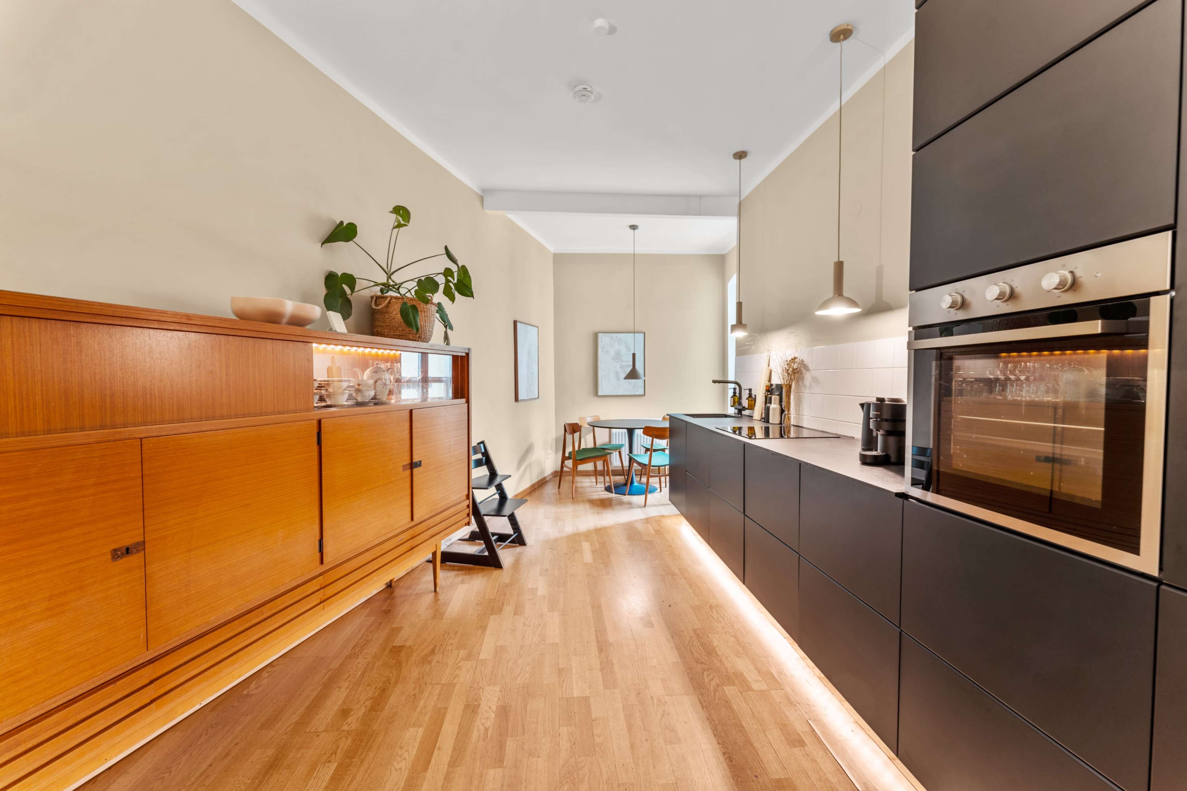 The image shows a modern kitchen featuring wooden cabinetry, black appliances, and a dining area with a small table and chairs.