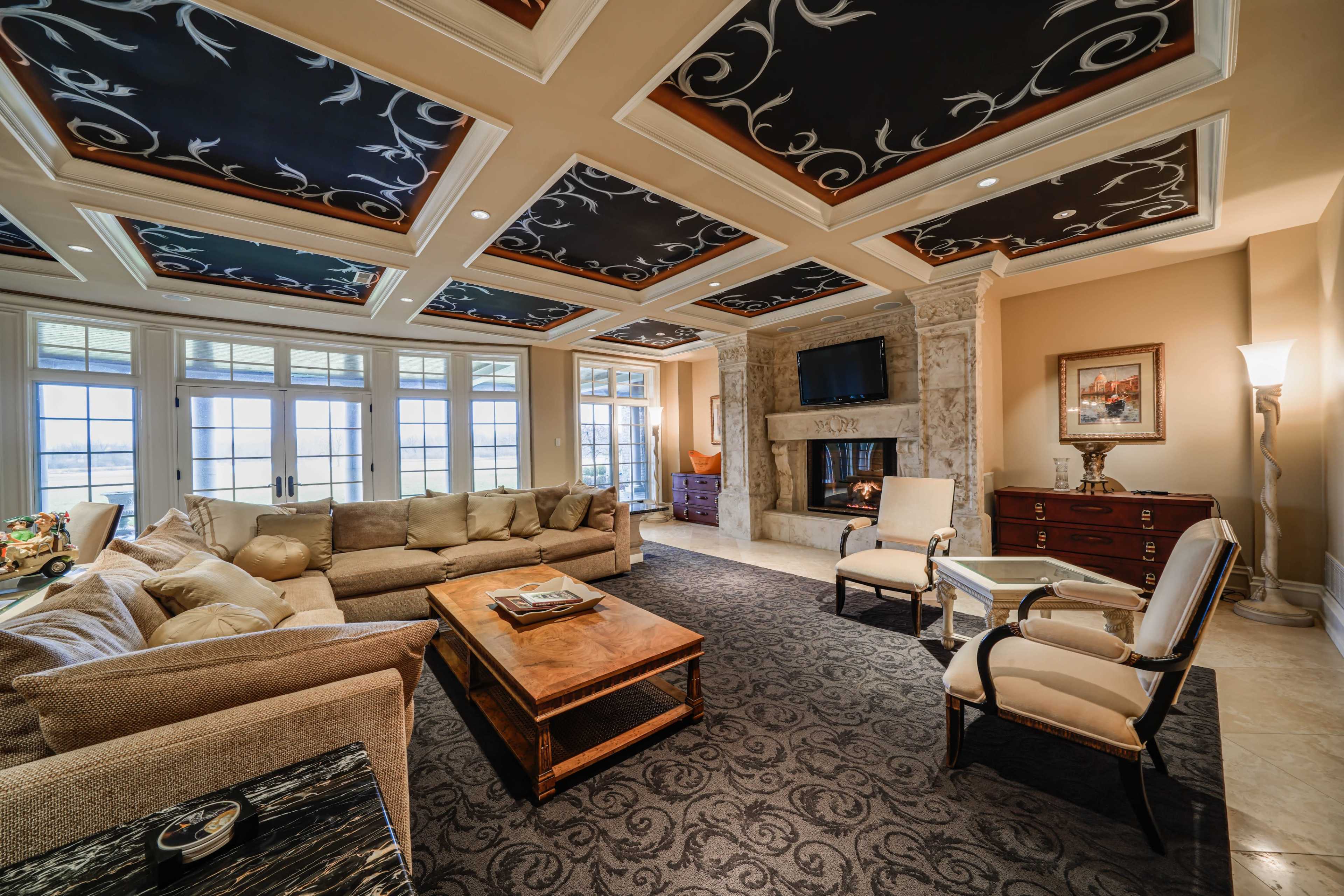The living room features a large sectional sofa, a wooden coffee table, and a stone fireplace, with decorative ceilings and multiple windows overlooking a view.