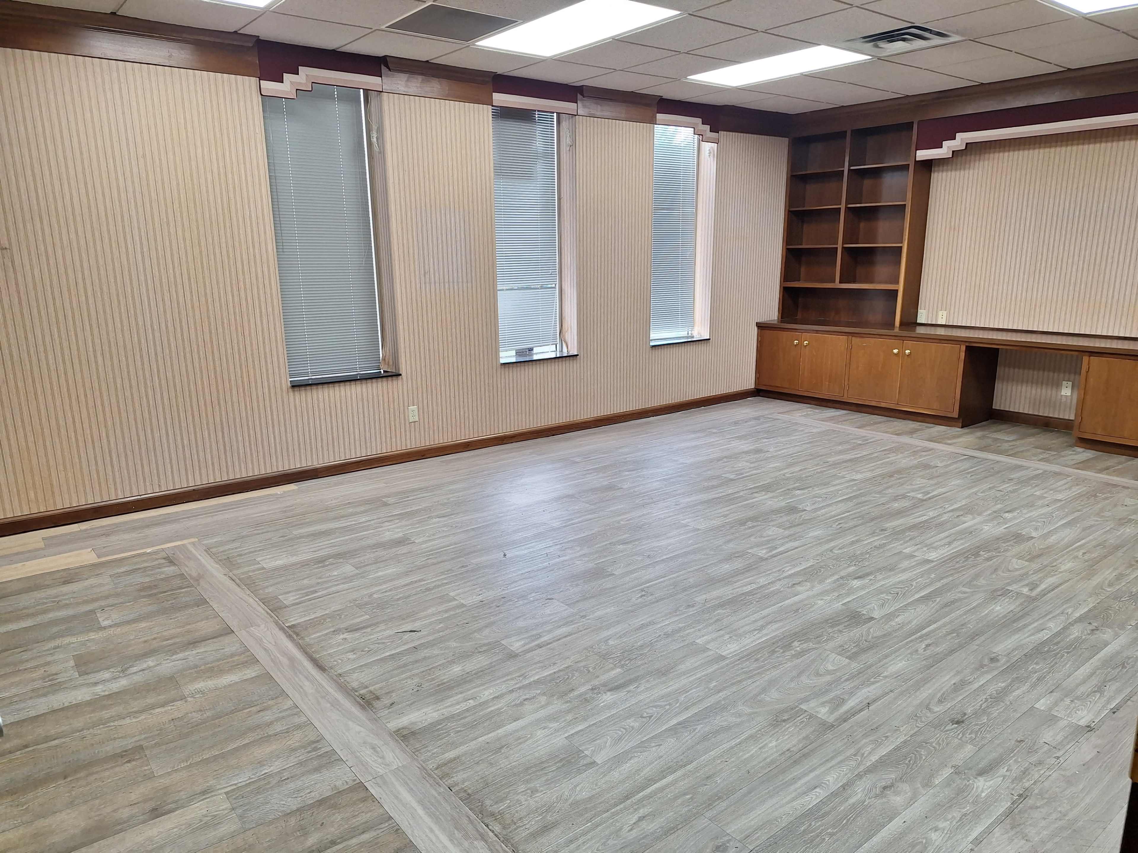 The image shows an empty room with wooden flooring, striped walls, tall windows, and a built-in shelving unit.