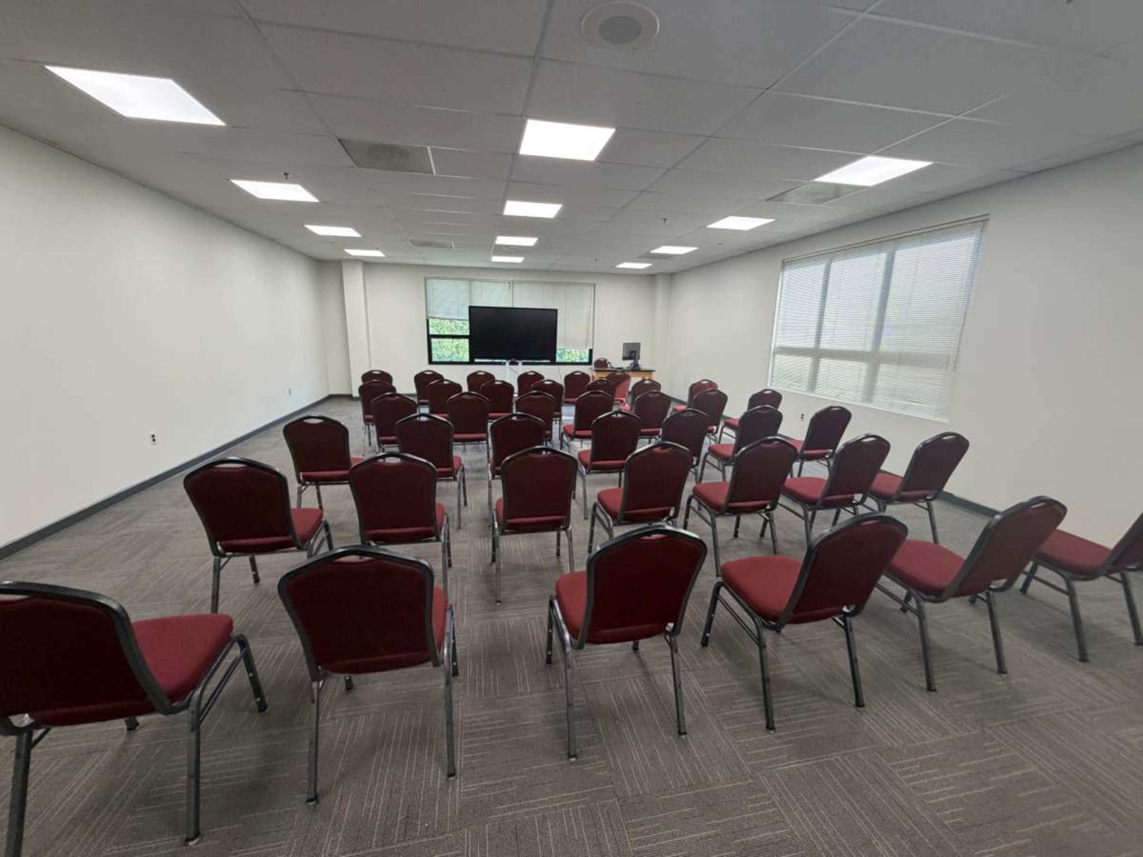 The image shows a conference room with rows of red chairs facing a large screen and a table at the front.