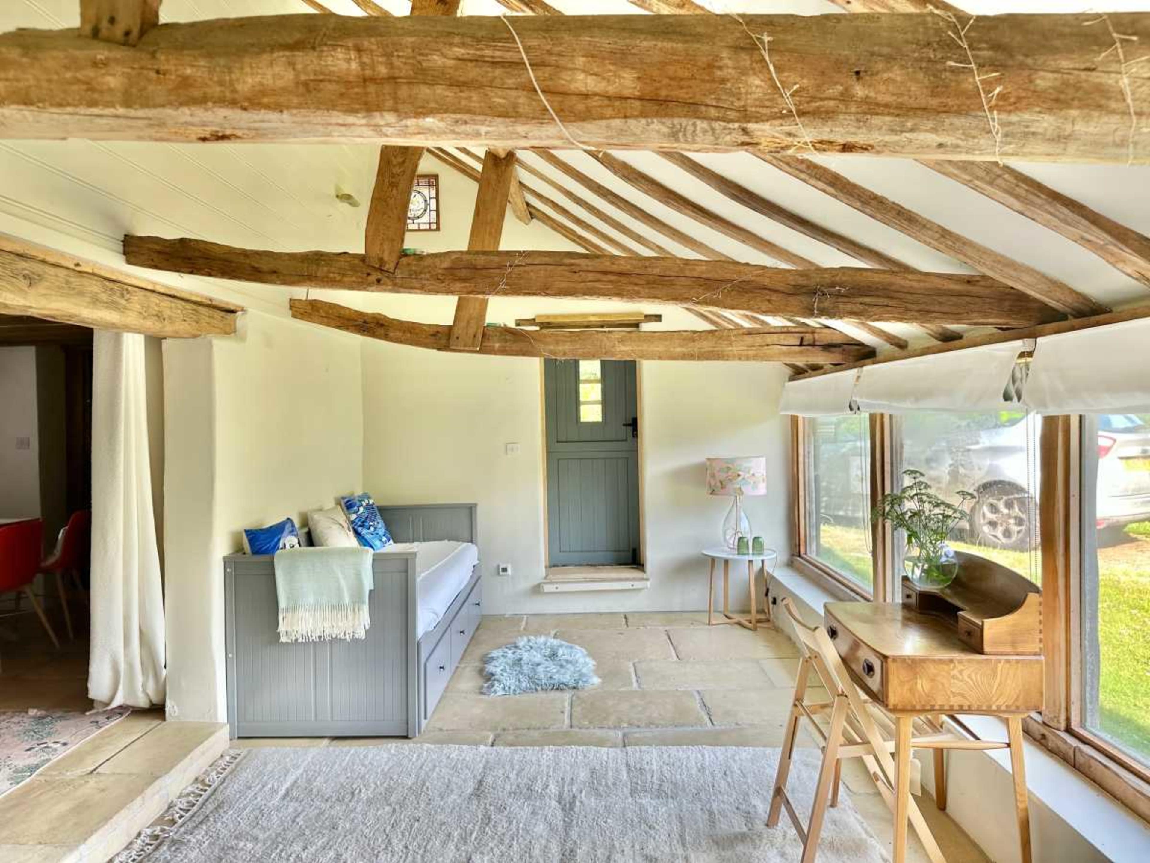 A cozy, light-filled living space features exposed wooden beams, a stone floor, and a combination of modern and rustic furnishings.