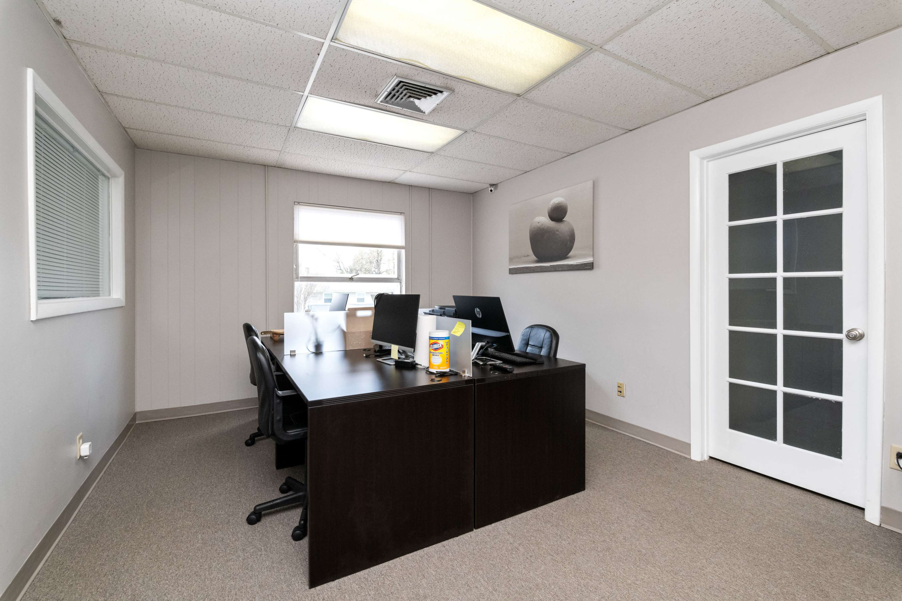 A sparsely furnished office with a large desk, two computer monitors, and a glass door leading to another room.