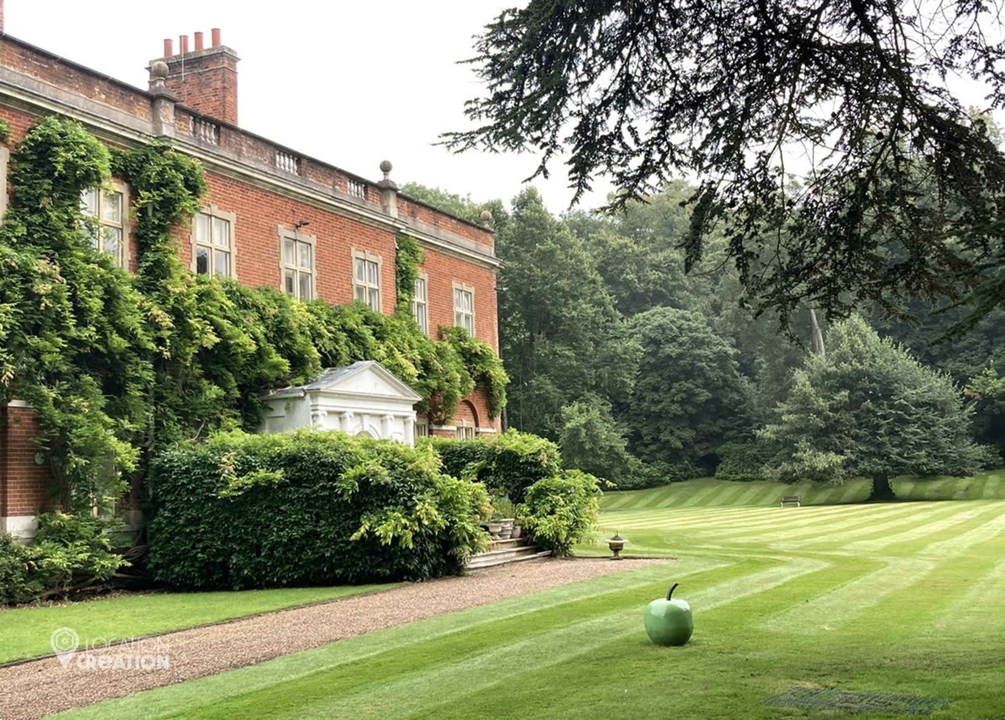 A large brick mansion covered in vines is surrounded by a neatly trimmed lawn featuring an oversized green apple sculpture.