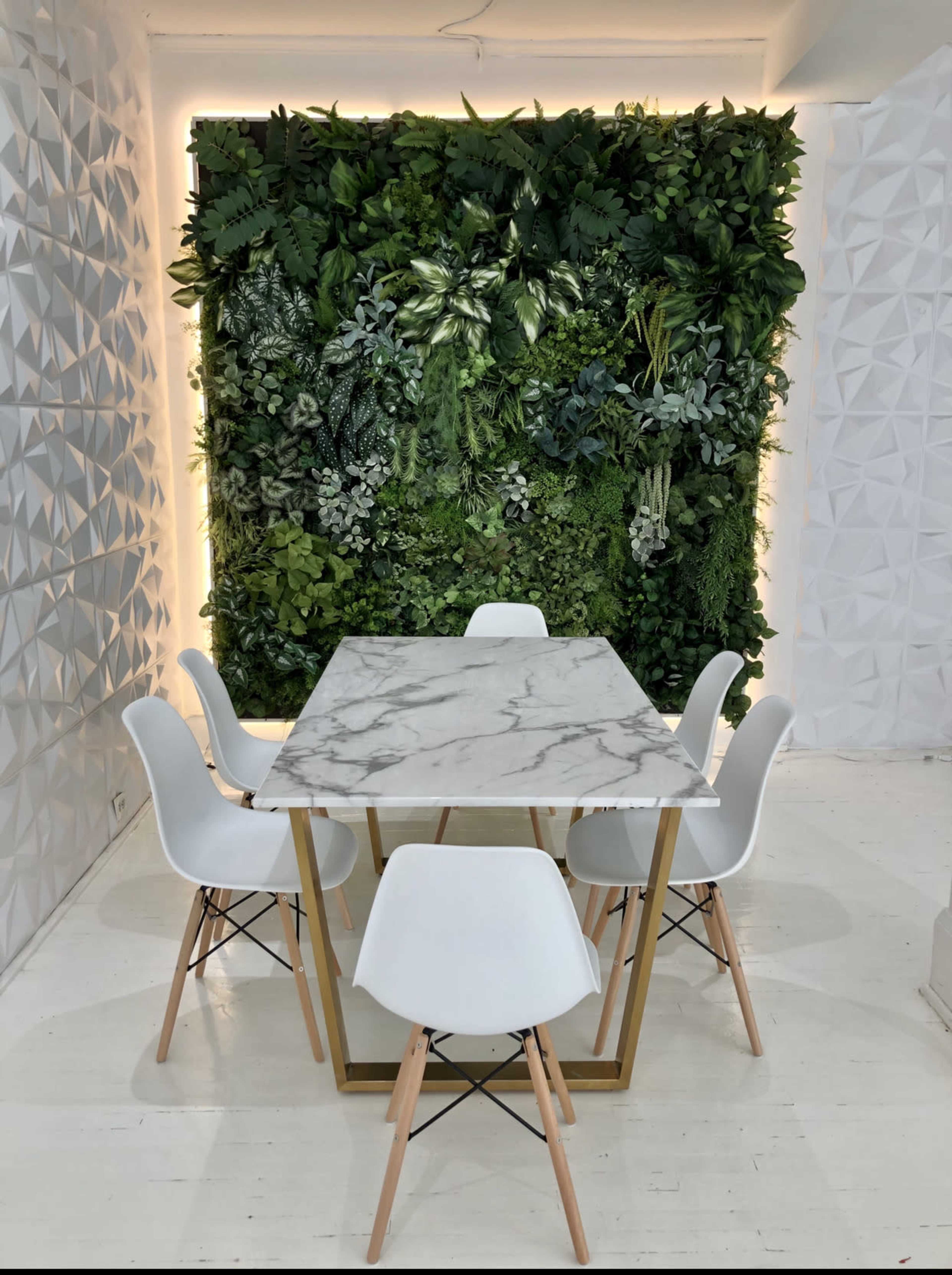 A marble-top dining table with six white chairs is set against a textured green wall composed of various plants.