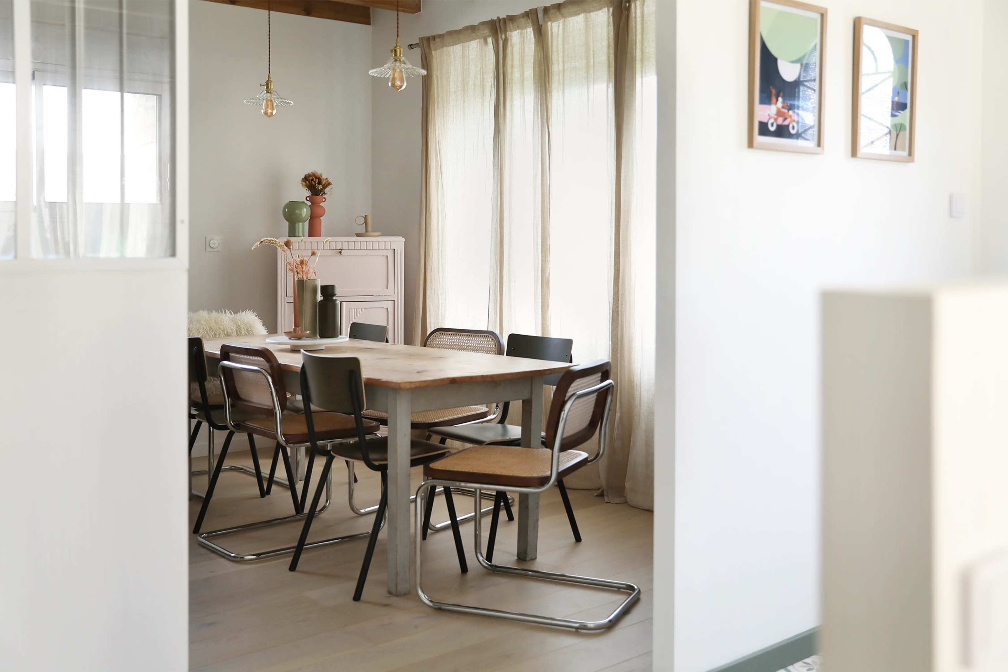 A dining room features a wooden table surrounded by metal and wicker chairs, with large windows and soft curtains allowing natural light to enter.
