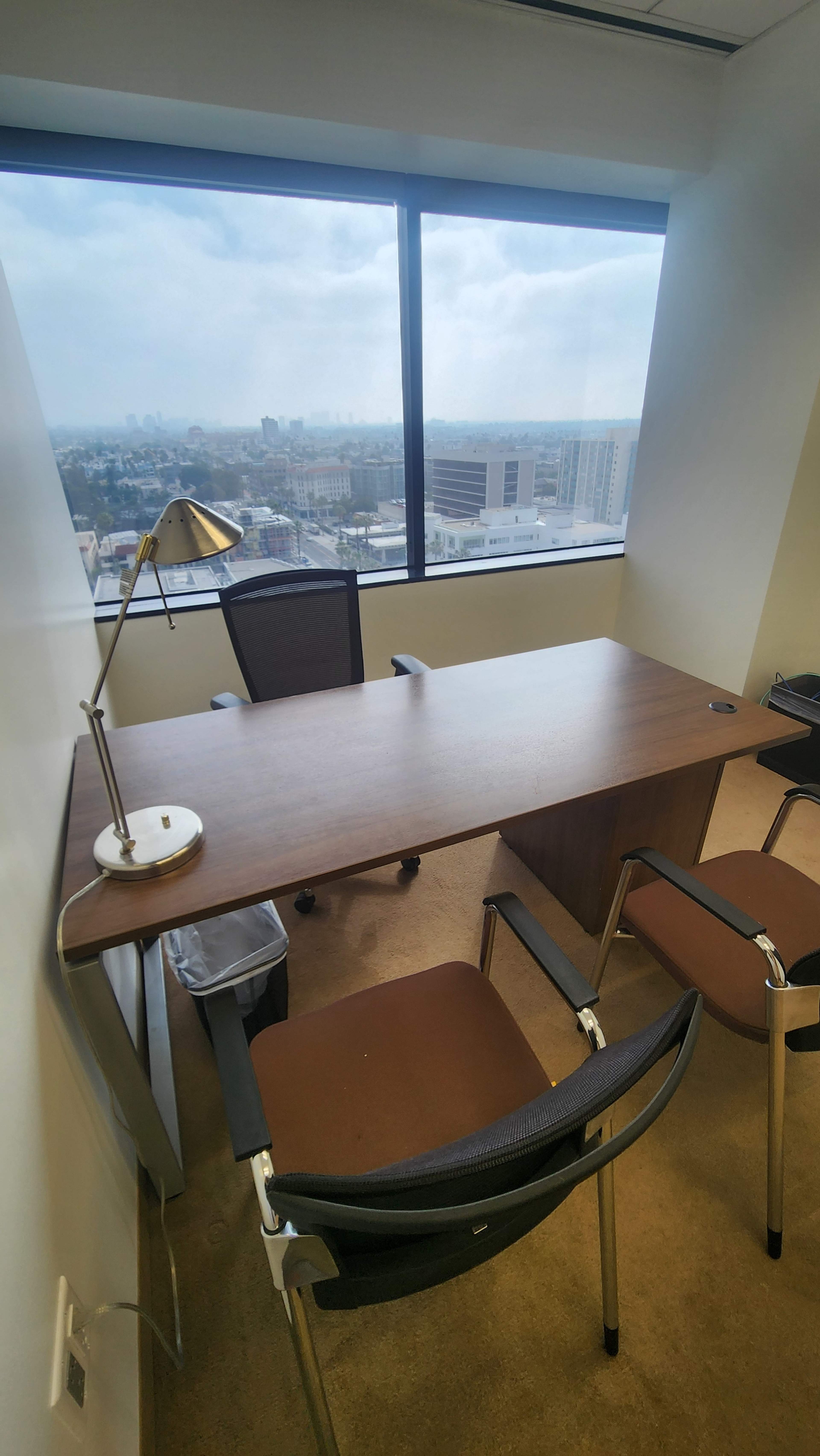 A sparsely furnished office featuring a wooden desk, two chairs, a desk lamp, and a large window with a view of the city skyline.