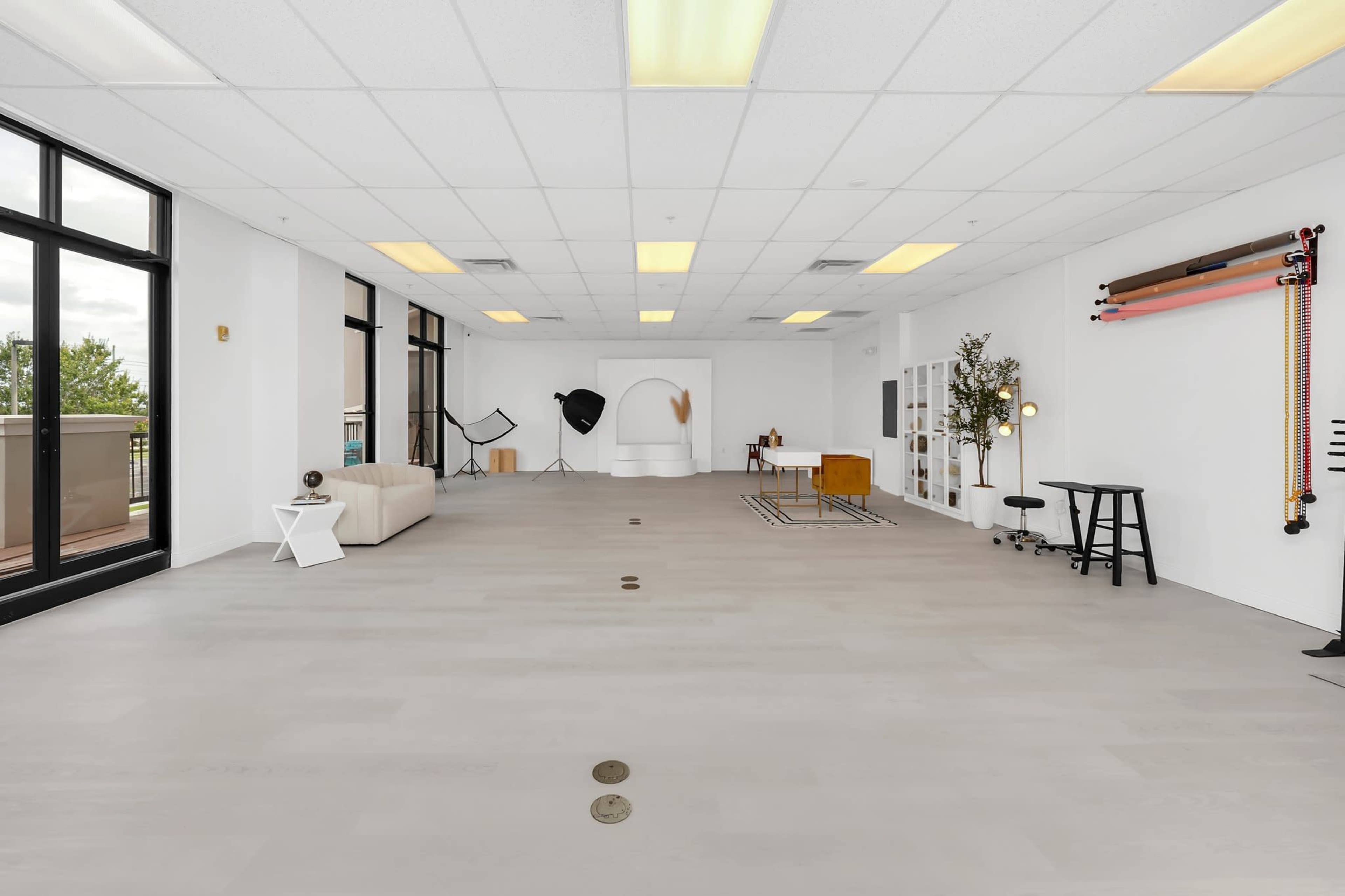 The image shows a spacious, minimalist room with large windows, featuring a sofa, a coffee table, and various modern decor elements scattered around.