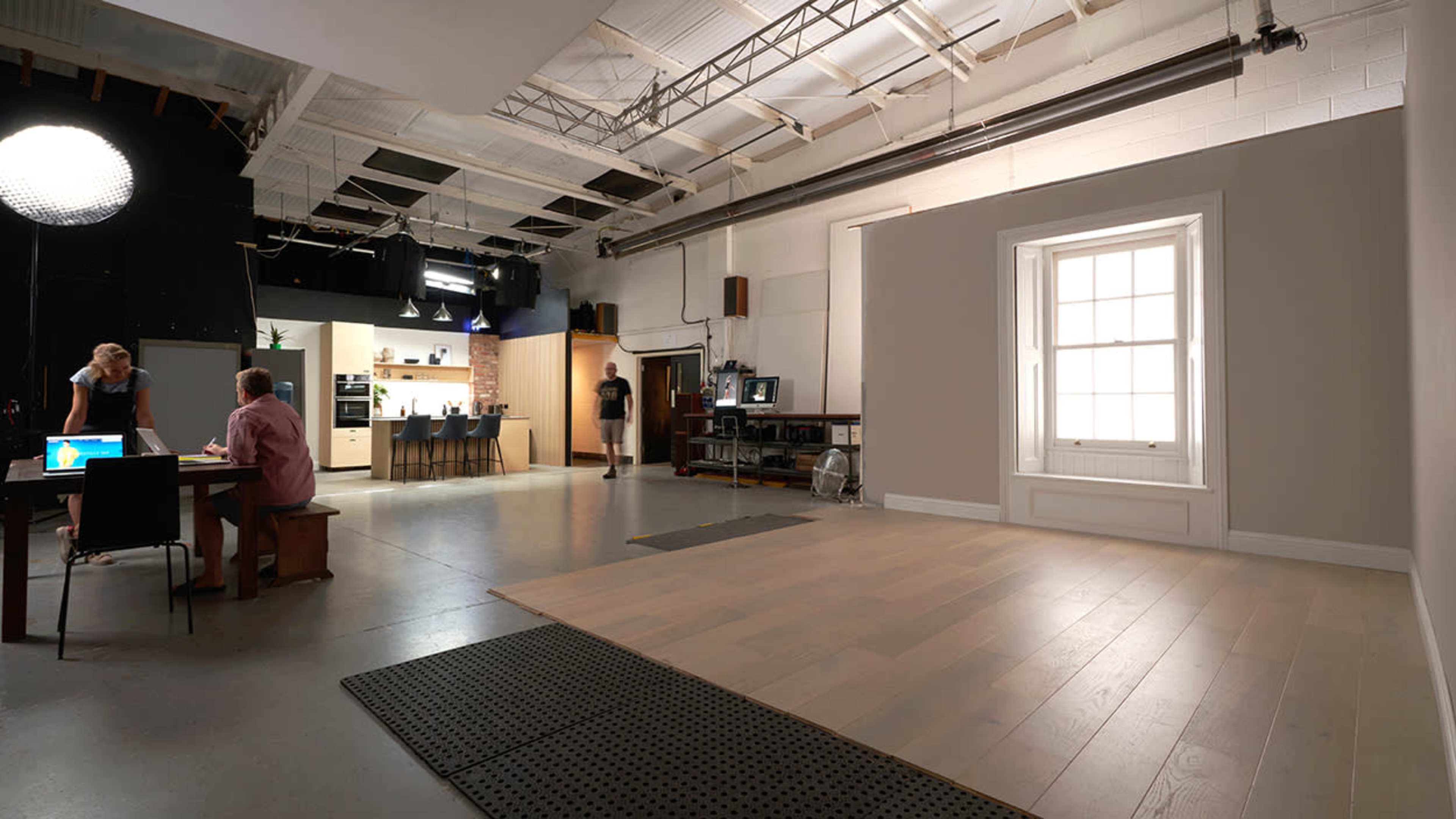 A spacious studio with a table and chairs on one side, while a well-lit kitchen area is visible in the background, and a large window is positioned along one wall.