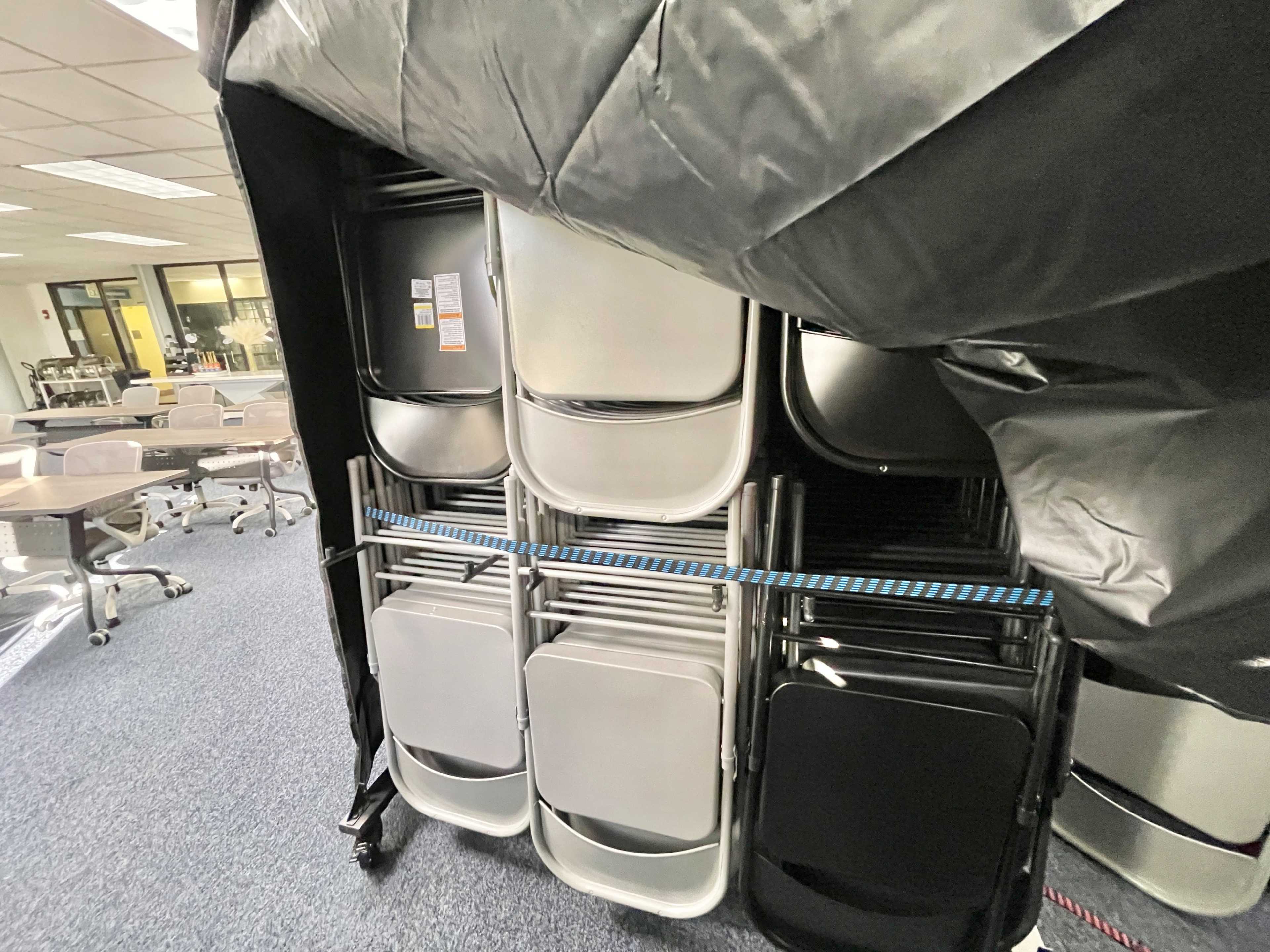 A stack of metal folding chairs is secured with a strap and covered by a black tarp in an office space.