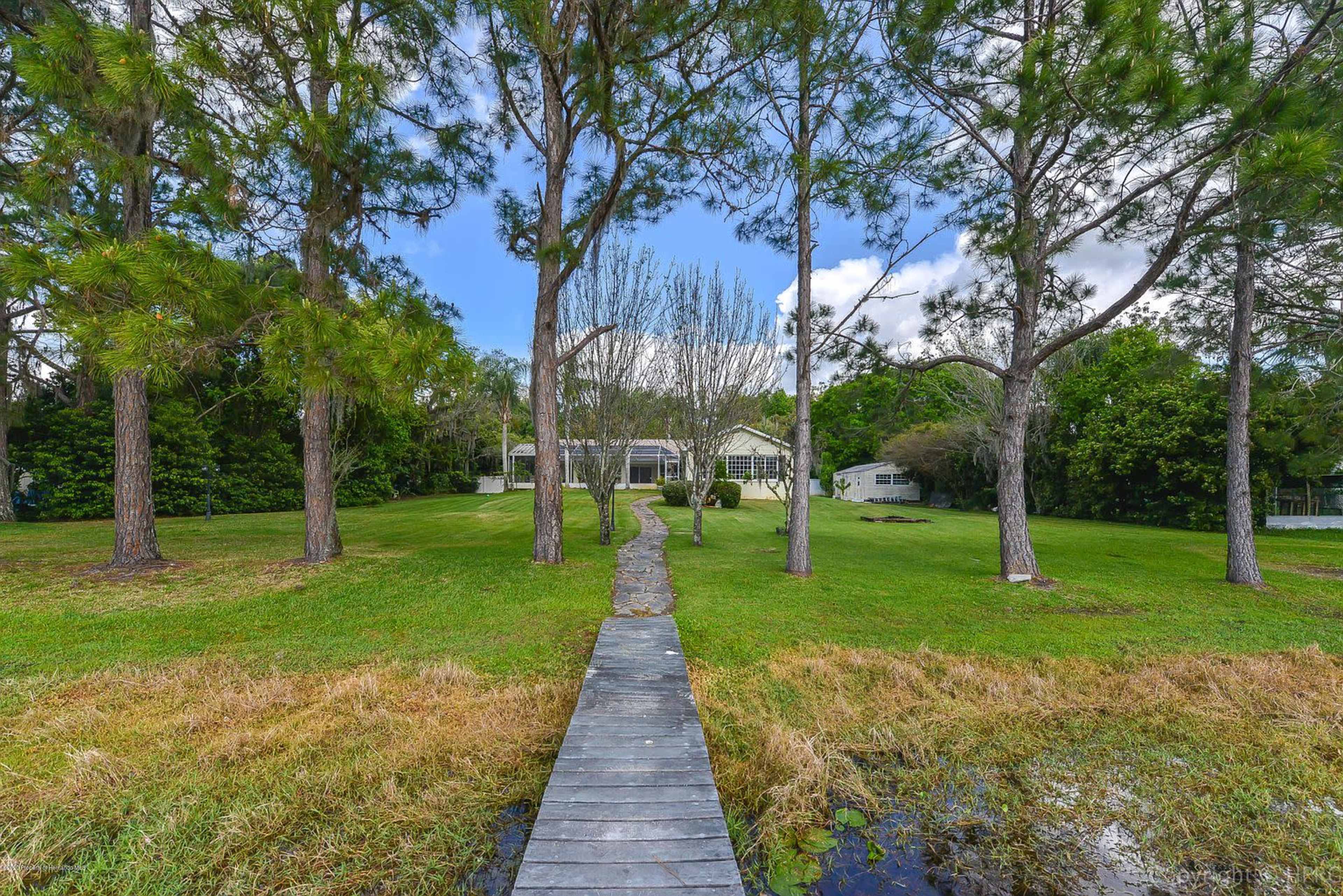 A wooden pathway leads from a body of water to a house surrounded by trees and grassy areas.