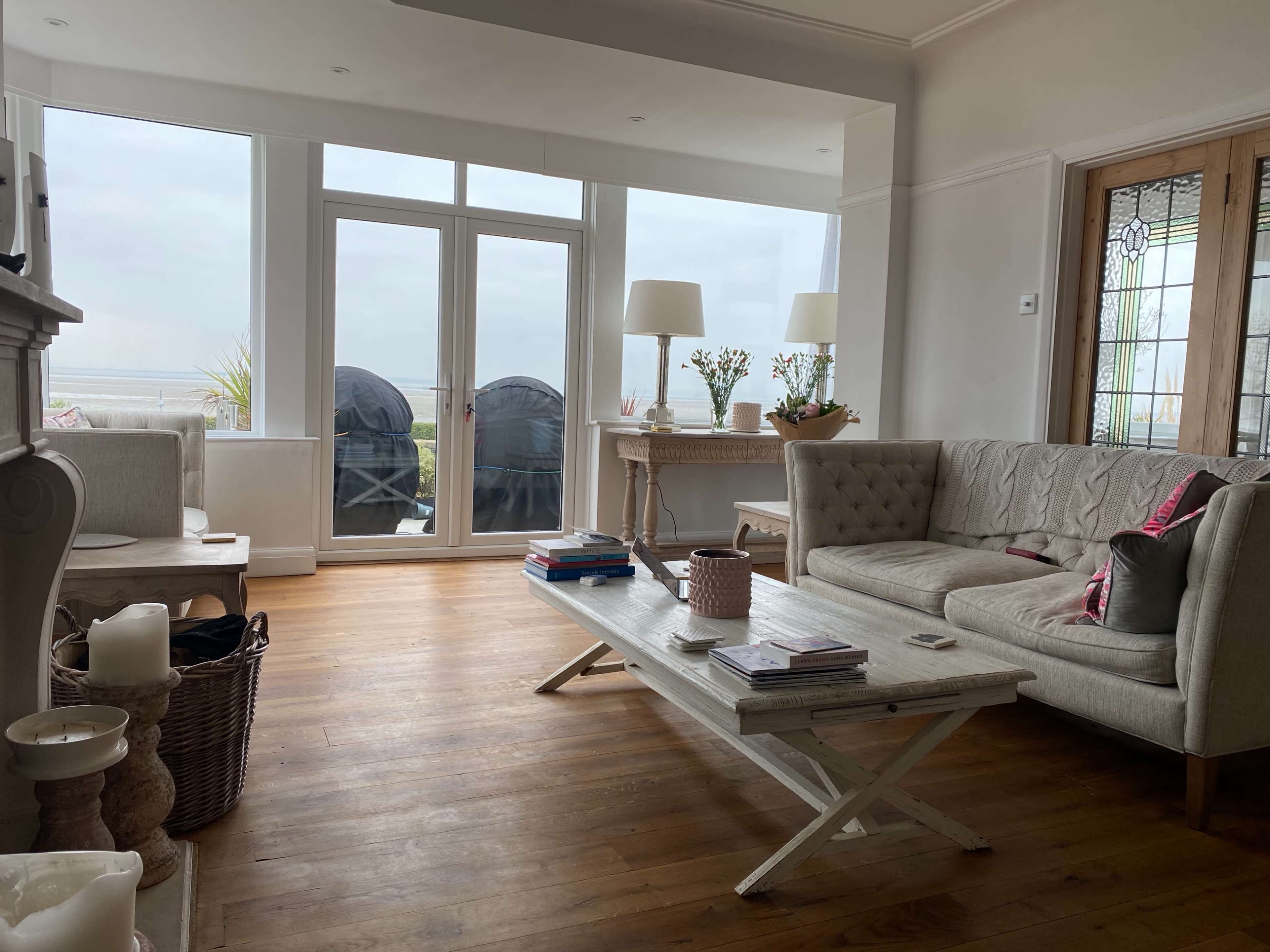 The living room features a large sofa, a coffee table stacked with magazines, and a view of the outdoors through large windows.