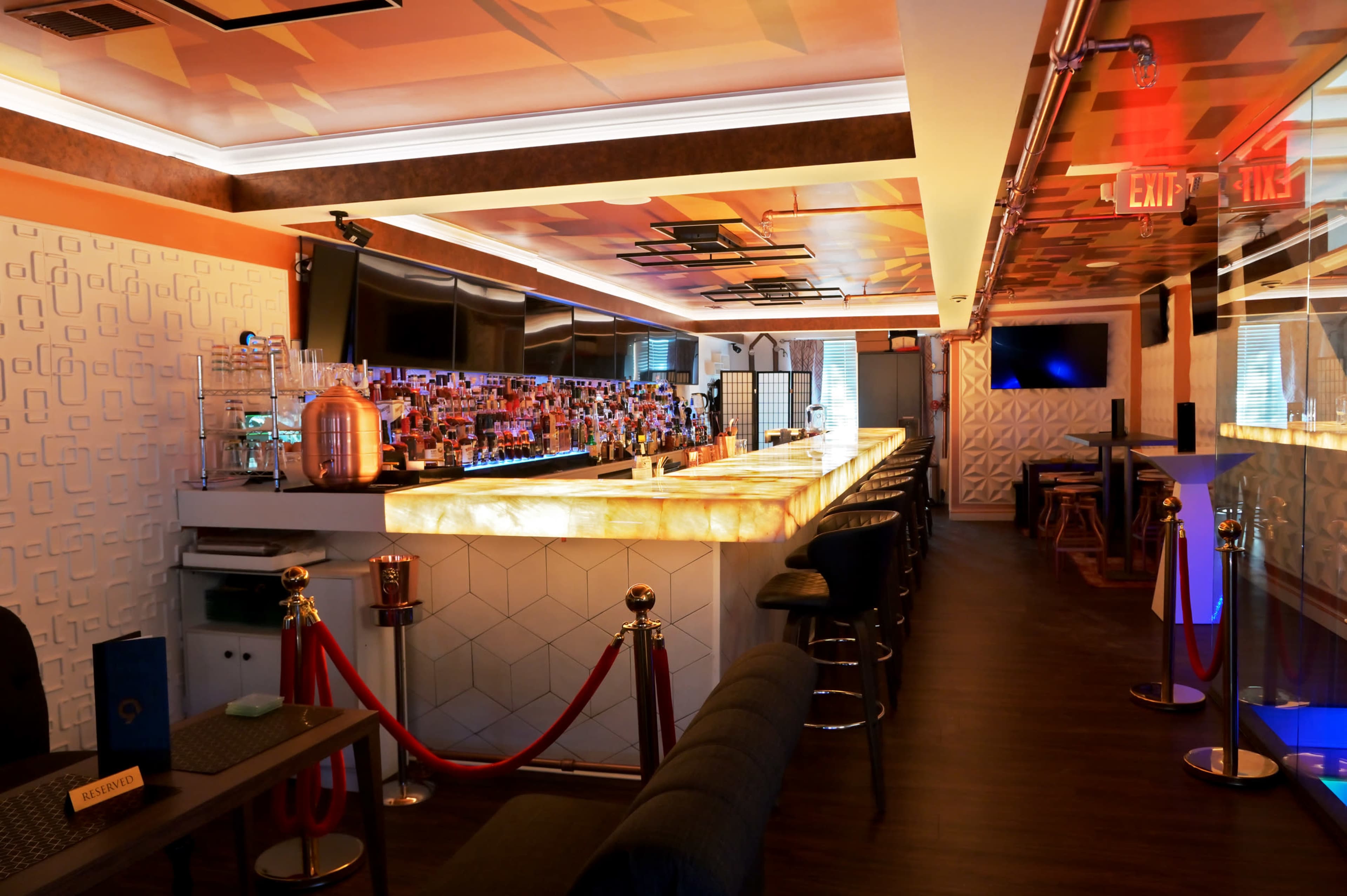 The image shows a stylish bar area featuring a backlit countertop, rows of liquor bottles, and a modern interior with a mix of seating arrangements.