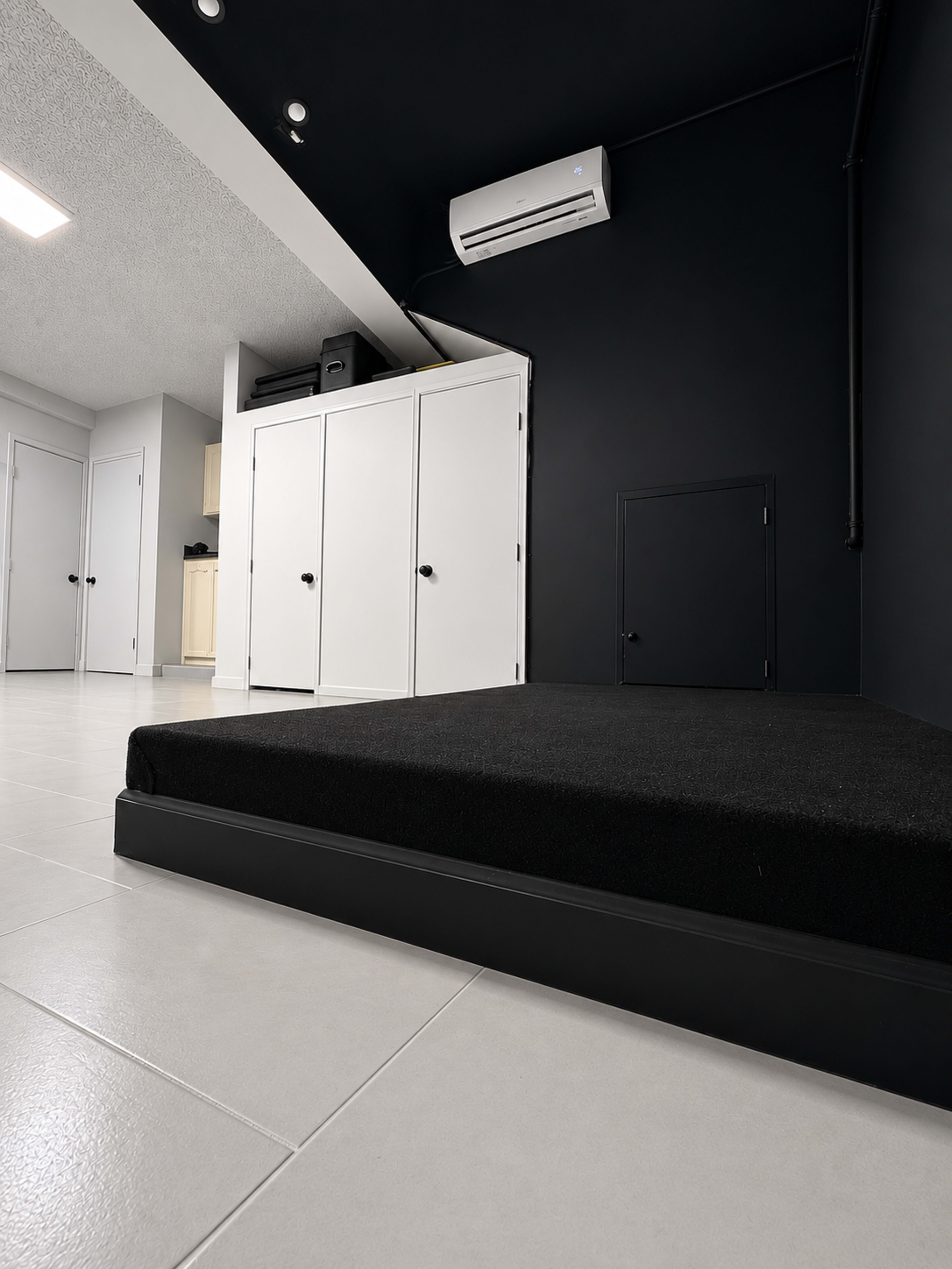 The image shows a minimalist room with a low black bed frame, polished white tiles, and black walls, featuring a small door and built-in white closets.
