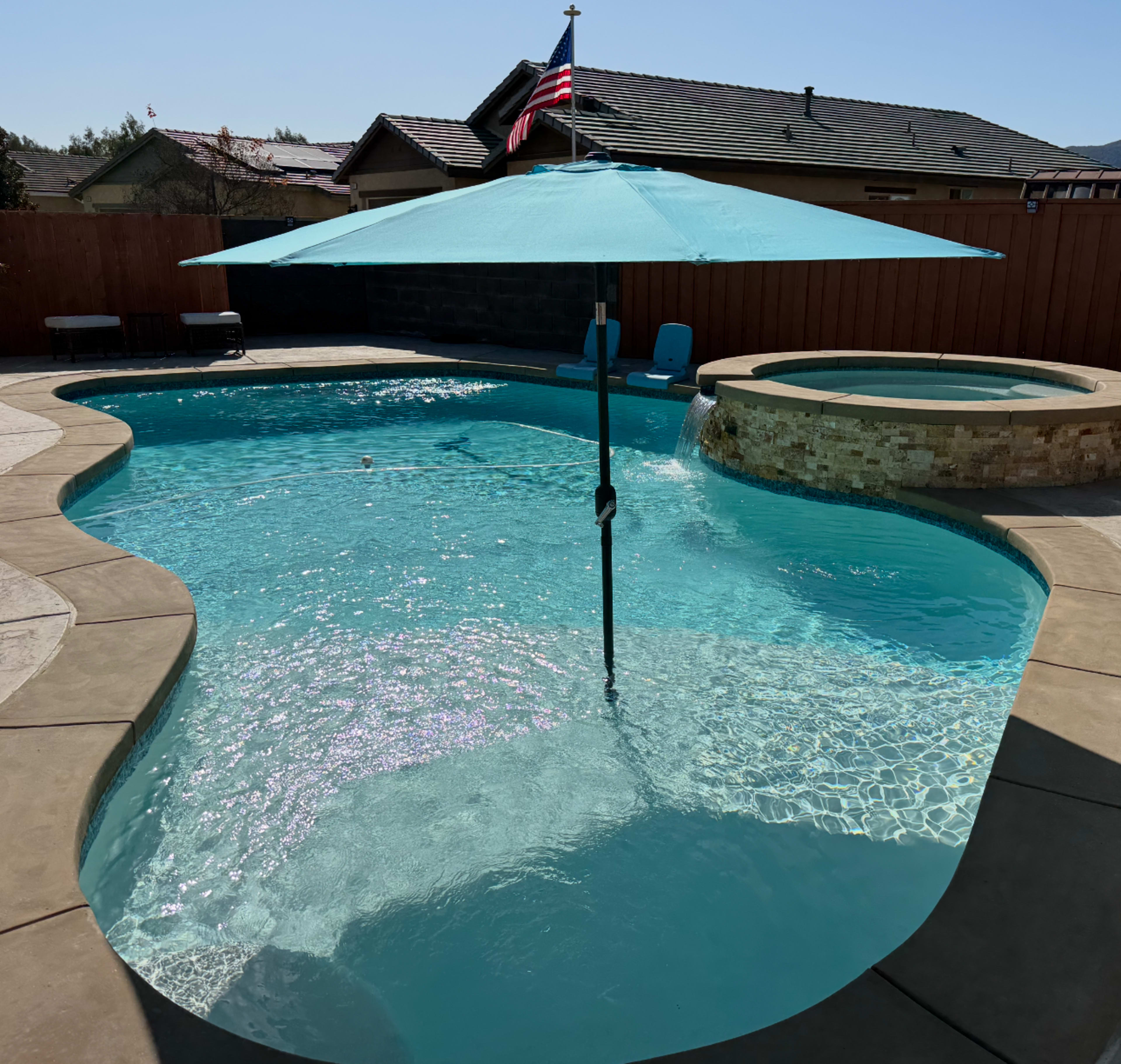 A turquoise umbrella shades a section of a swimming pool surrounded by a stone patio and a raised hot tub.