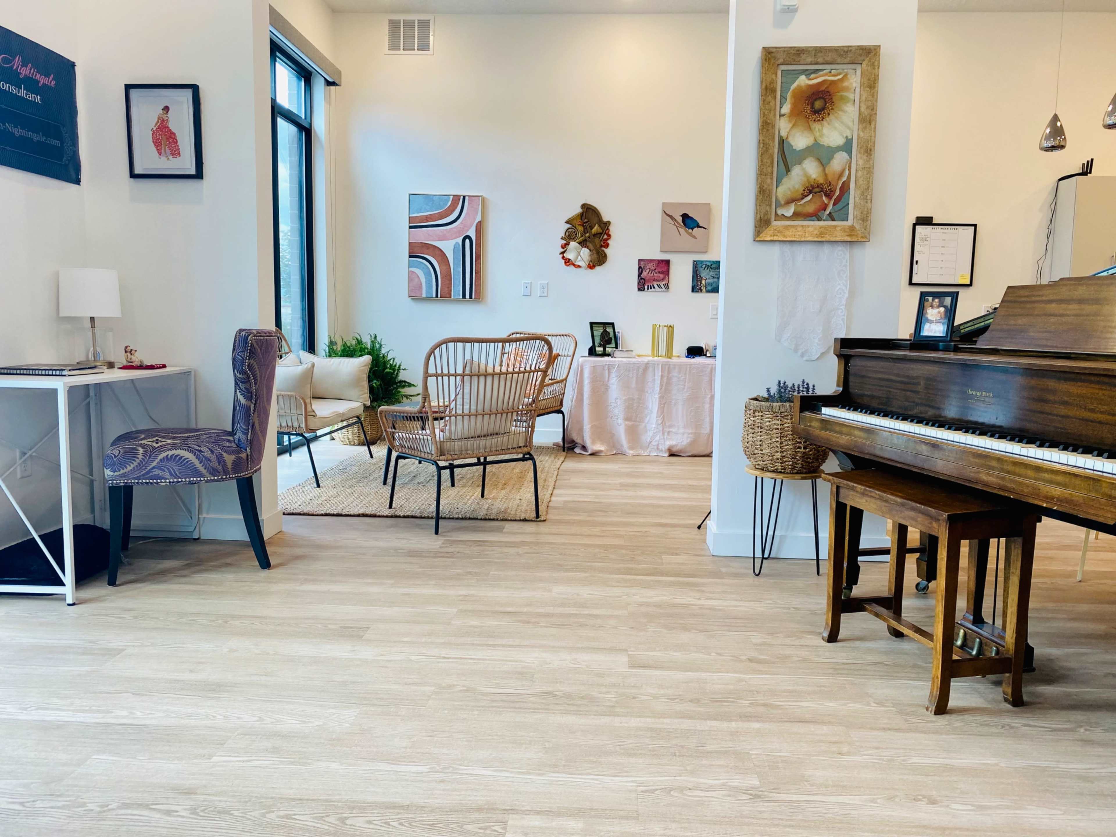 The image shows a bright, modern interior featuring a grand piano, a small desk with a chair, and a seating area with various artworks on the walls.