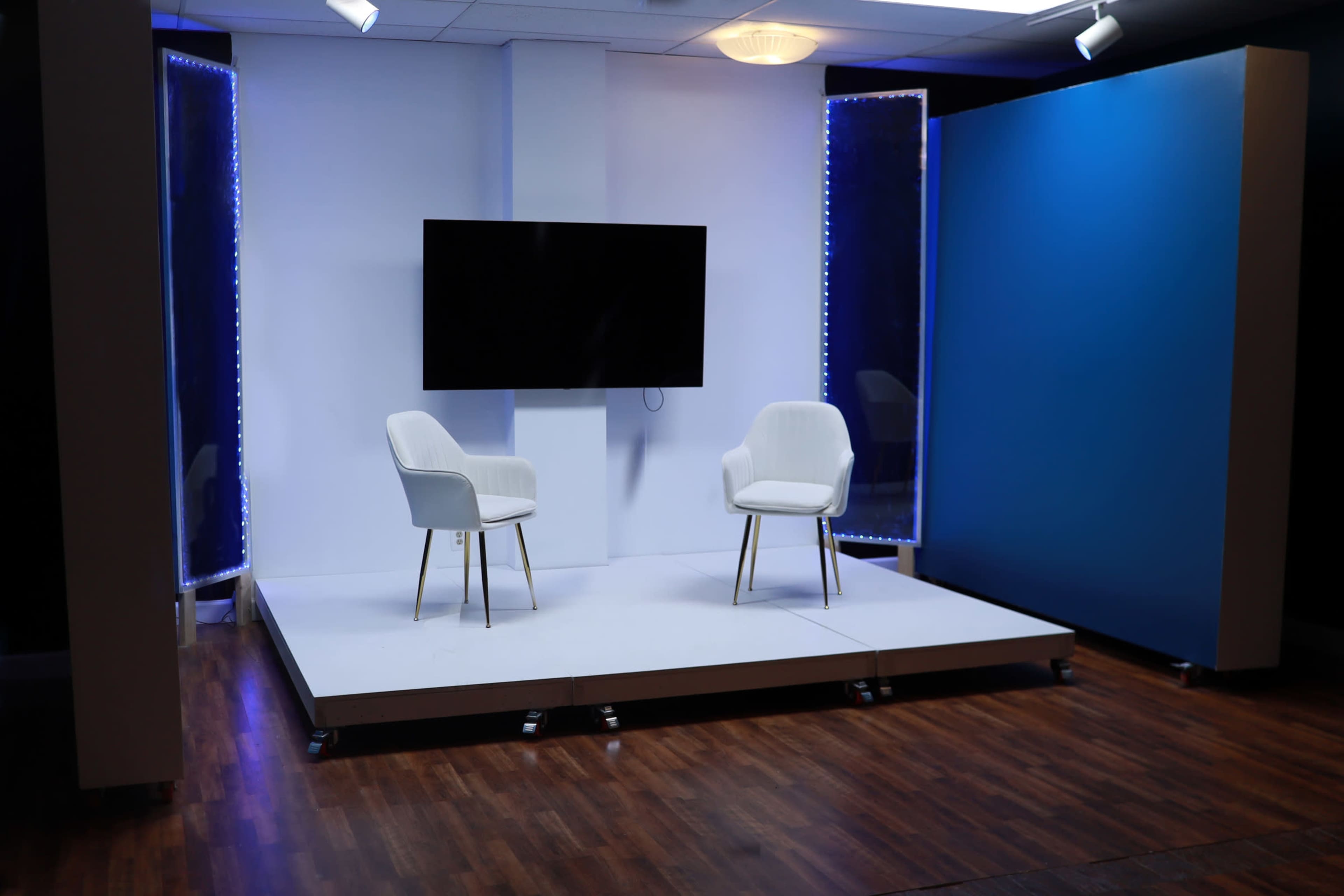 A minimalist studio setup features two white chairs on a raised platform with a large screen and blue backdrop.