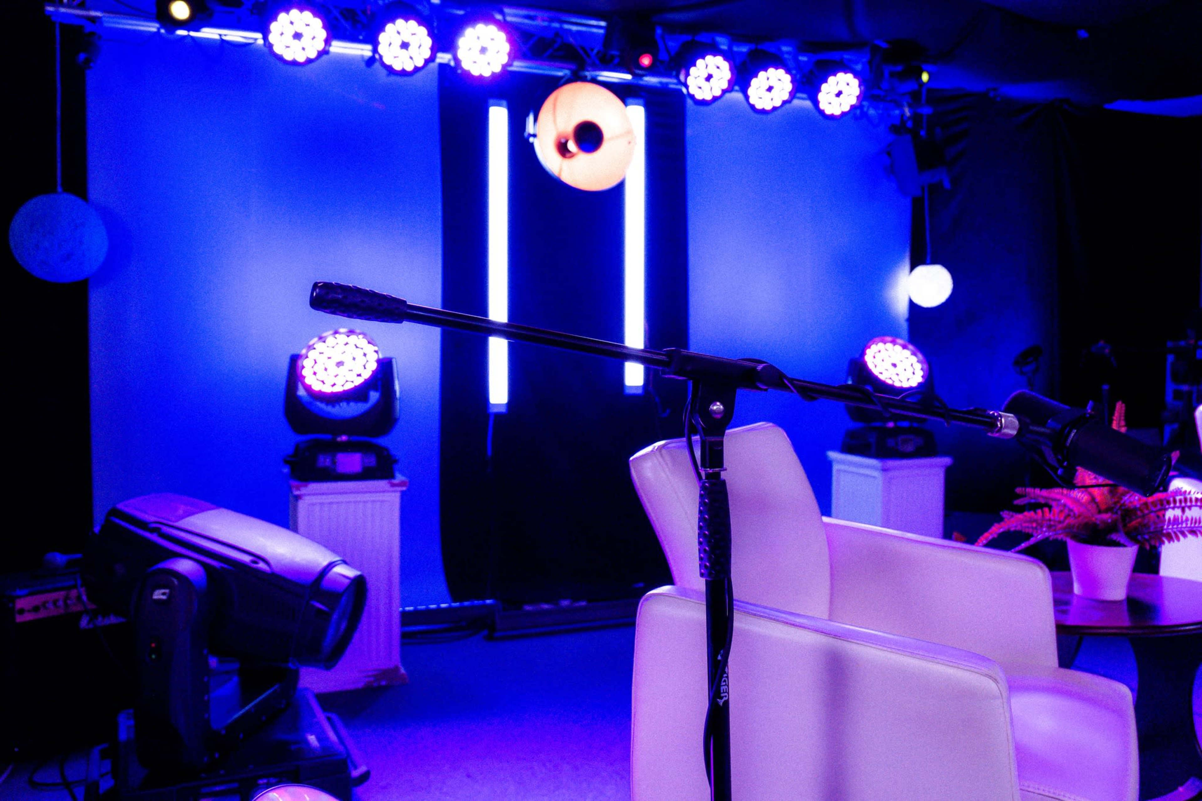 The image shows a stage setup with a microphone, a white chair, and colorful lighting featuring purple hues.
