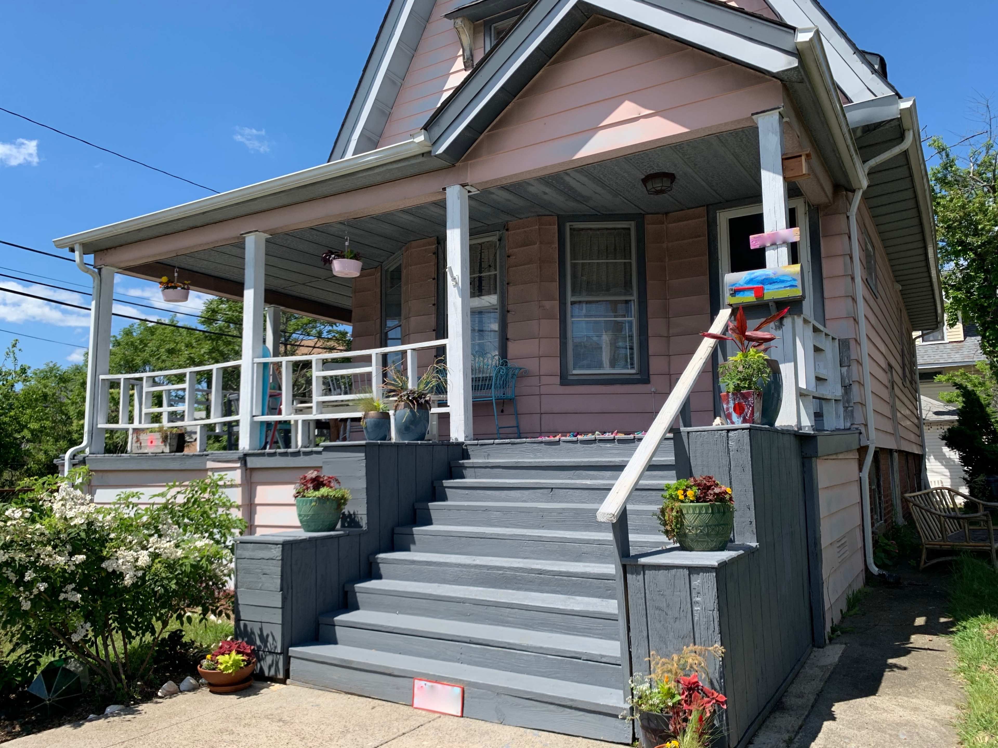 The image shows a pink two-story house with a porch, featuring decorated steps and potted plants along the entrance.