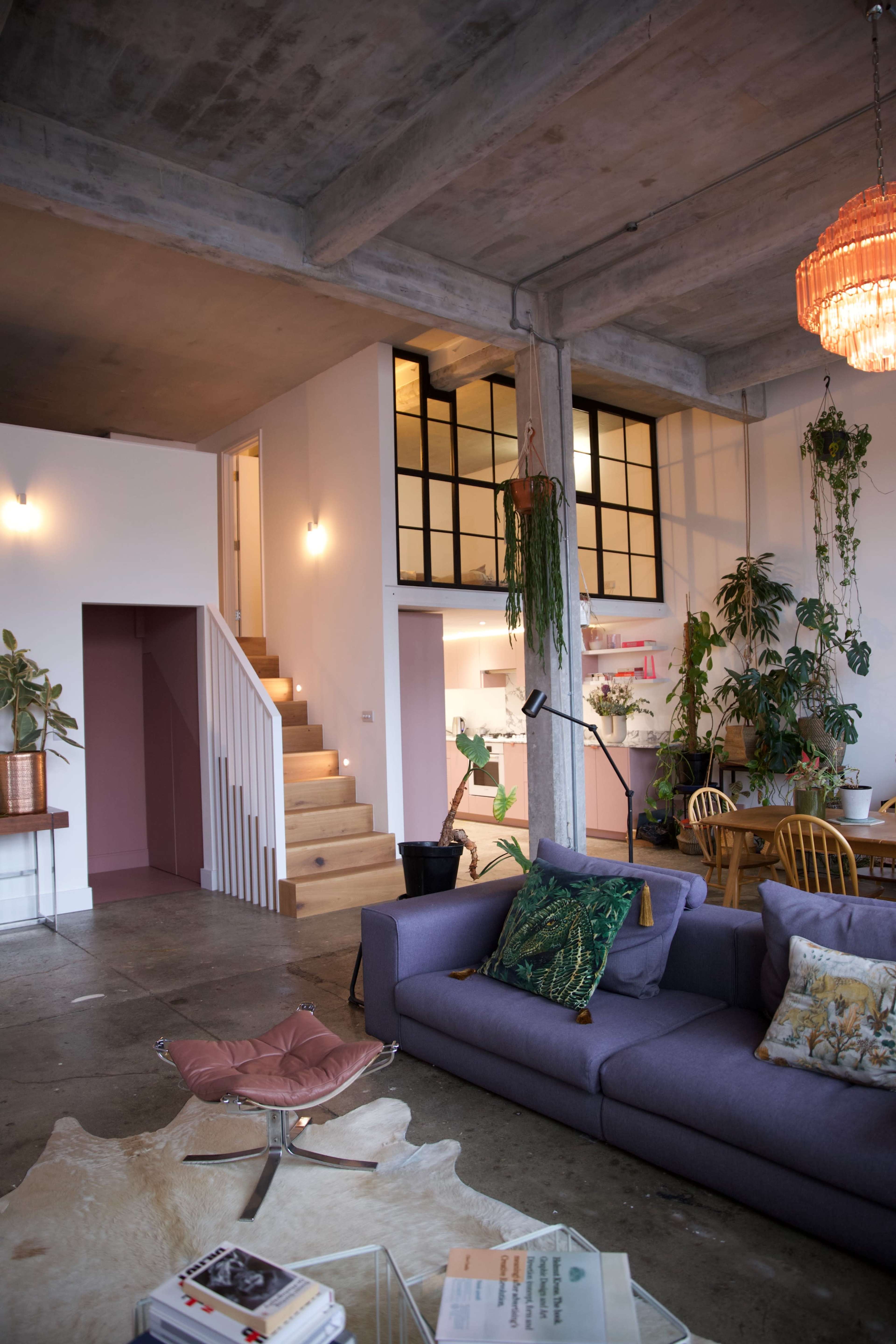 The image shows a modern living space with a purple sofa, a dining area, and a staircase leading to an upstairs section surrounded by indoor plants.