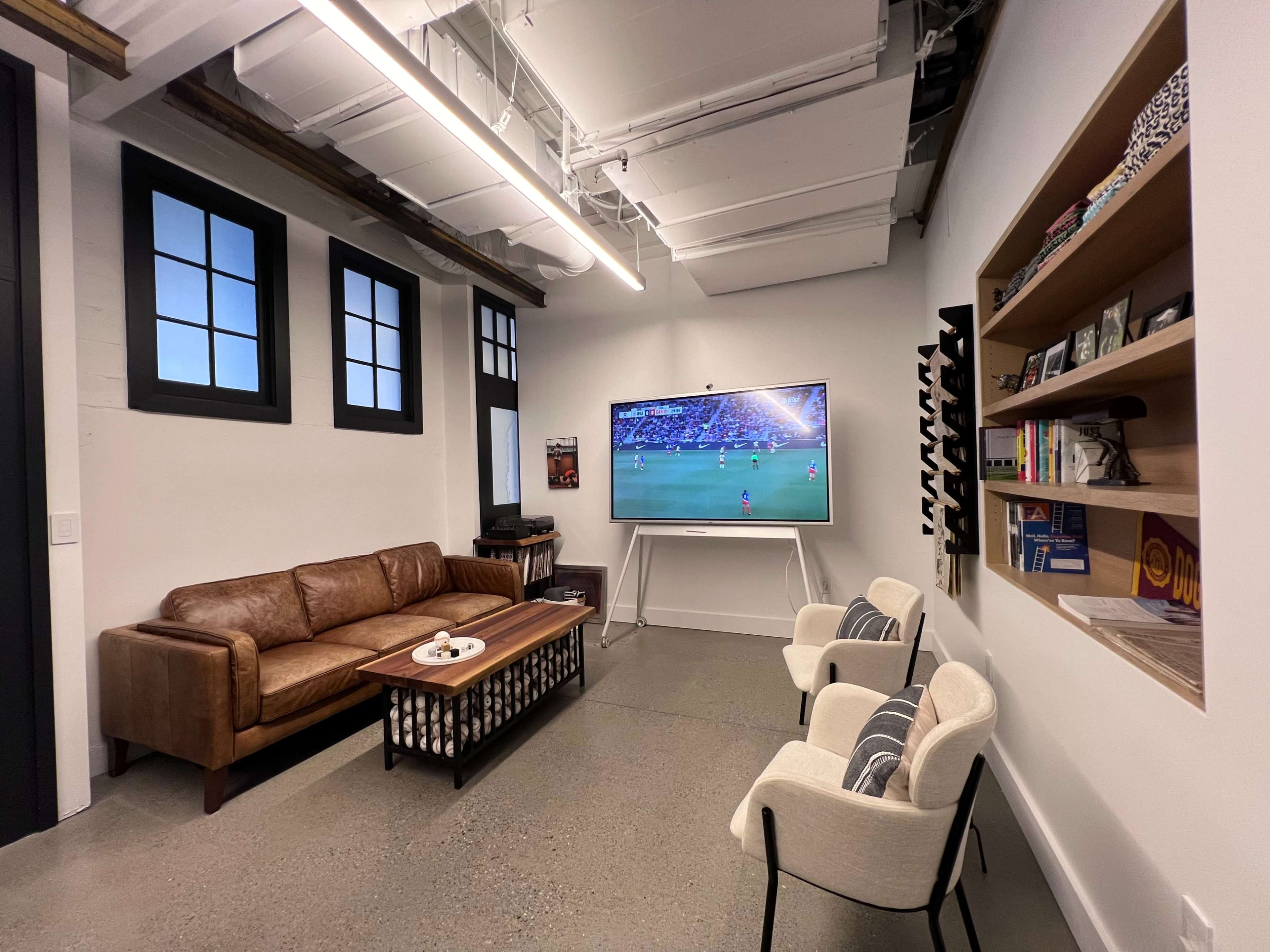 A modern lounge area with a brown leather sofa, two chairs, a coffee table, and a large television displaying a sports event, along with shelves containing books and decorative items.