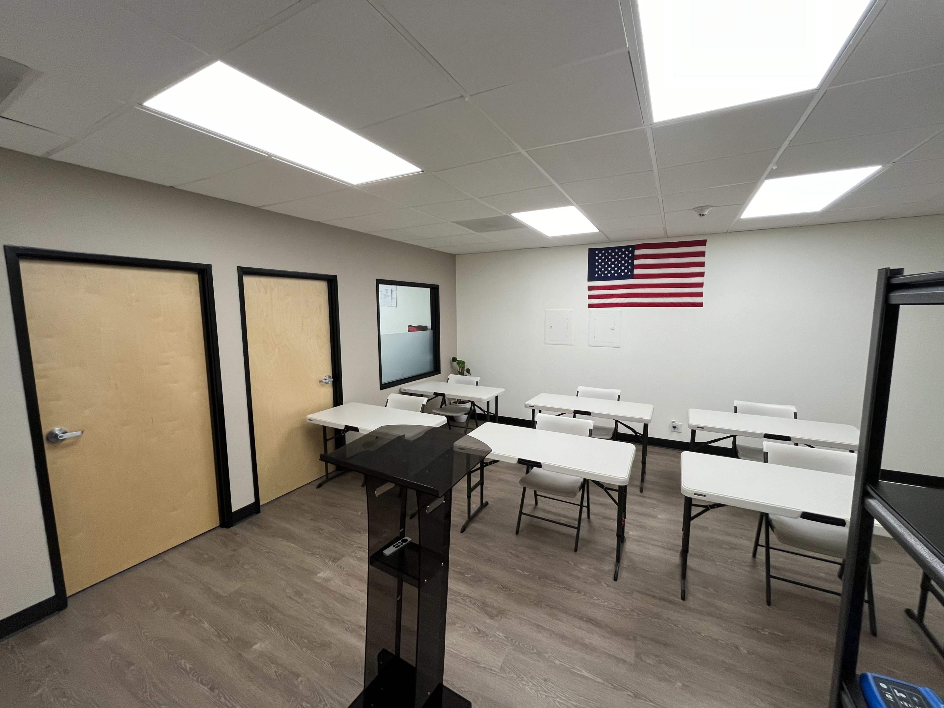 A classroom with white desks arranged in rows, a podium at the front, and an American flag hanging on the wall.