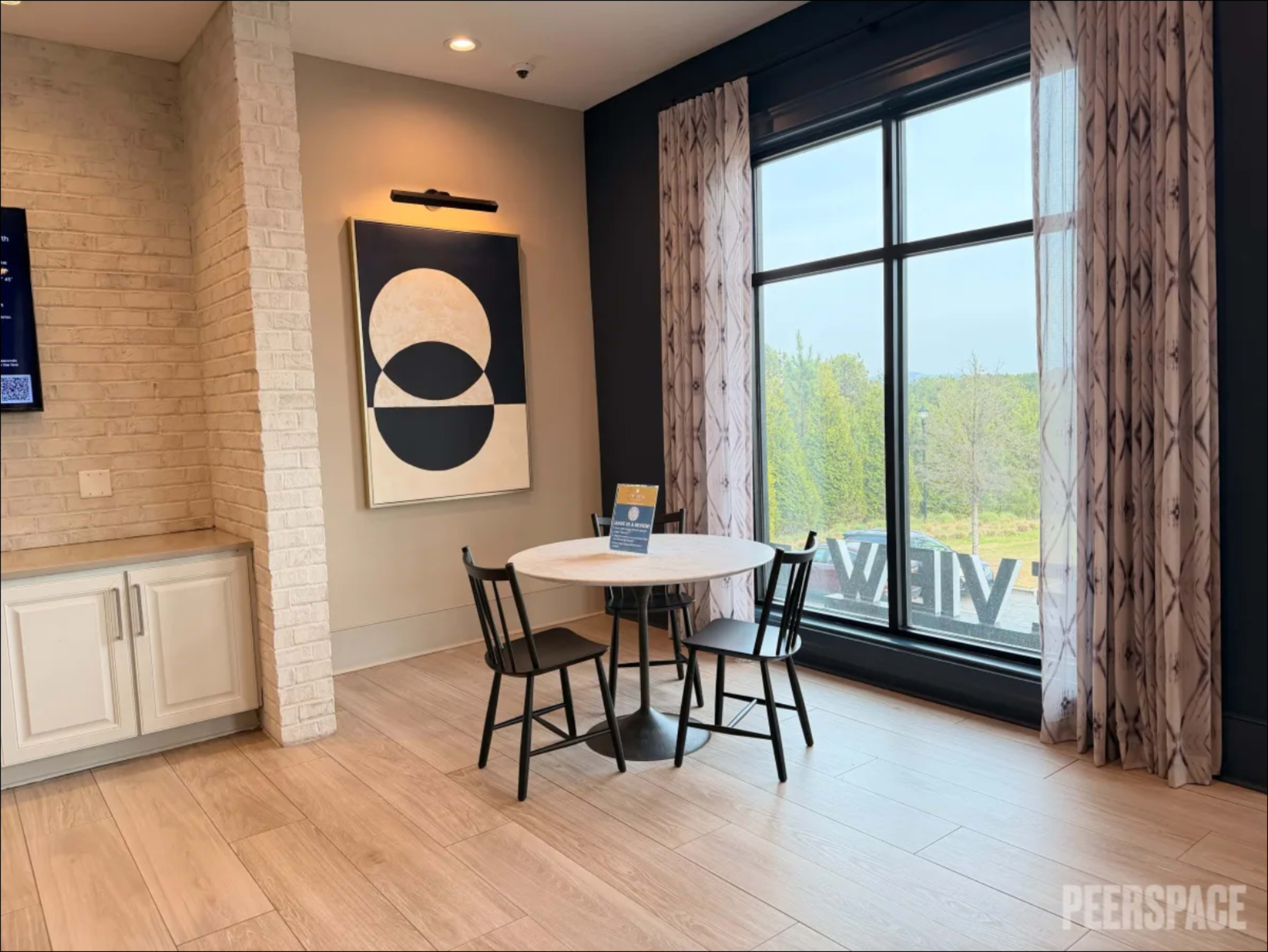 A round table with two black chairs is set by a large window overlooking green trees, with a modern artwork displayed on the wall nearby.