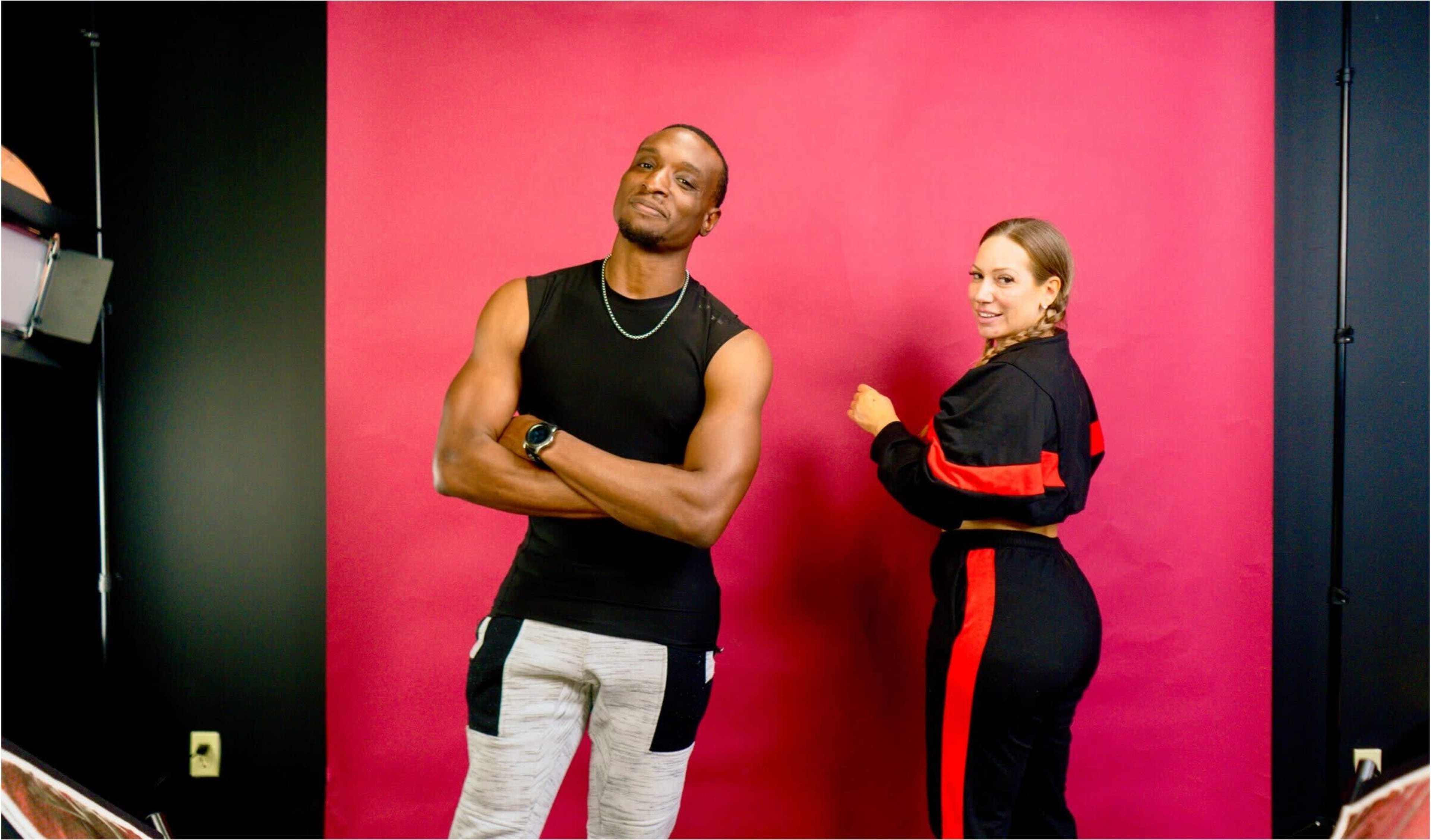 A man with muscular arms stands confidently in front of a pink backdrop, while a woman in athletic wear poses with her back turned.
