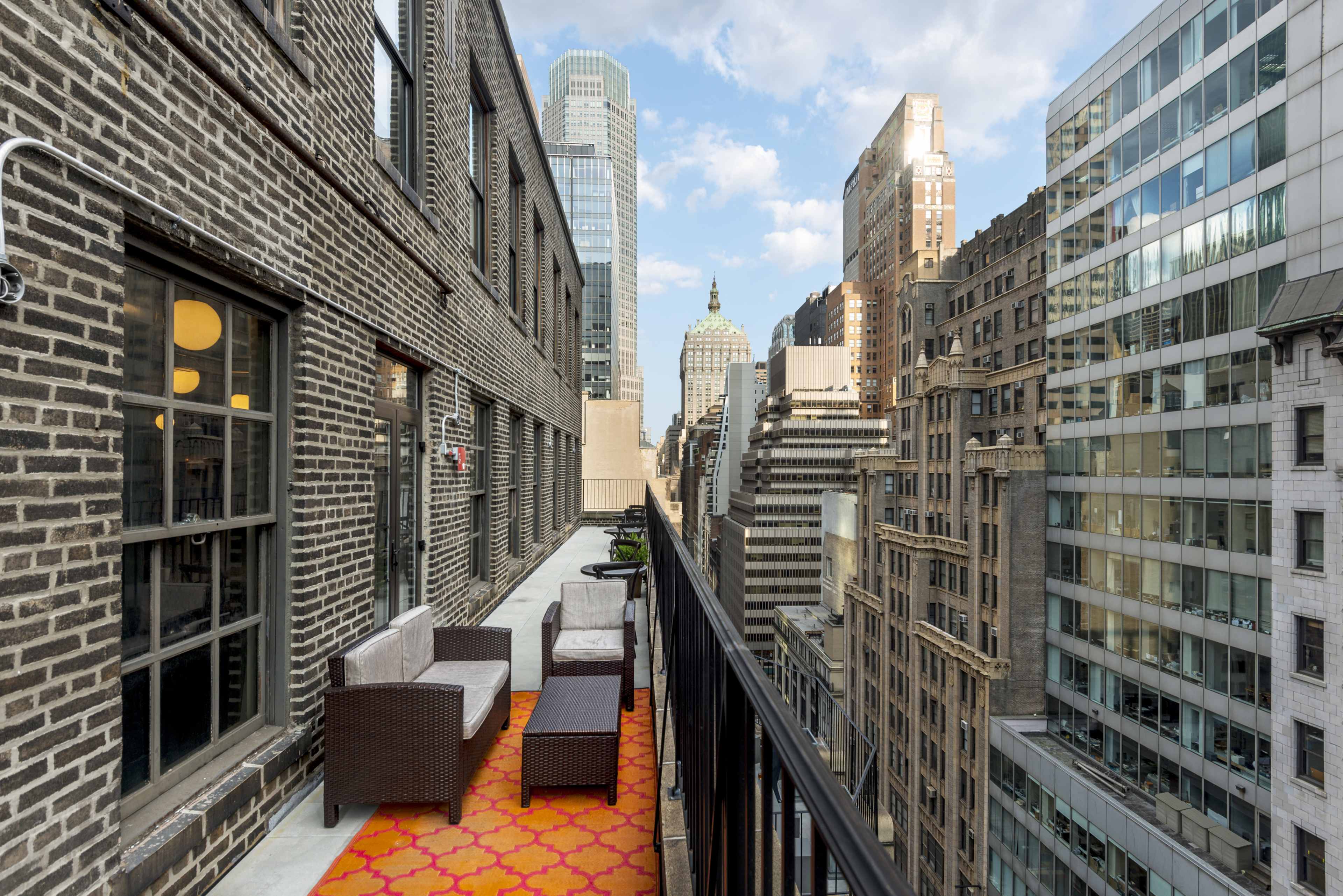 The image shows a balcony with two chairs and an orange patterned rug, overlooking a city skyline featuring a mix of modern and historic buildings under a partly cloudy sky.