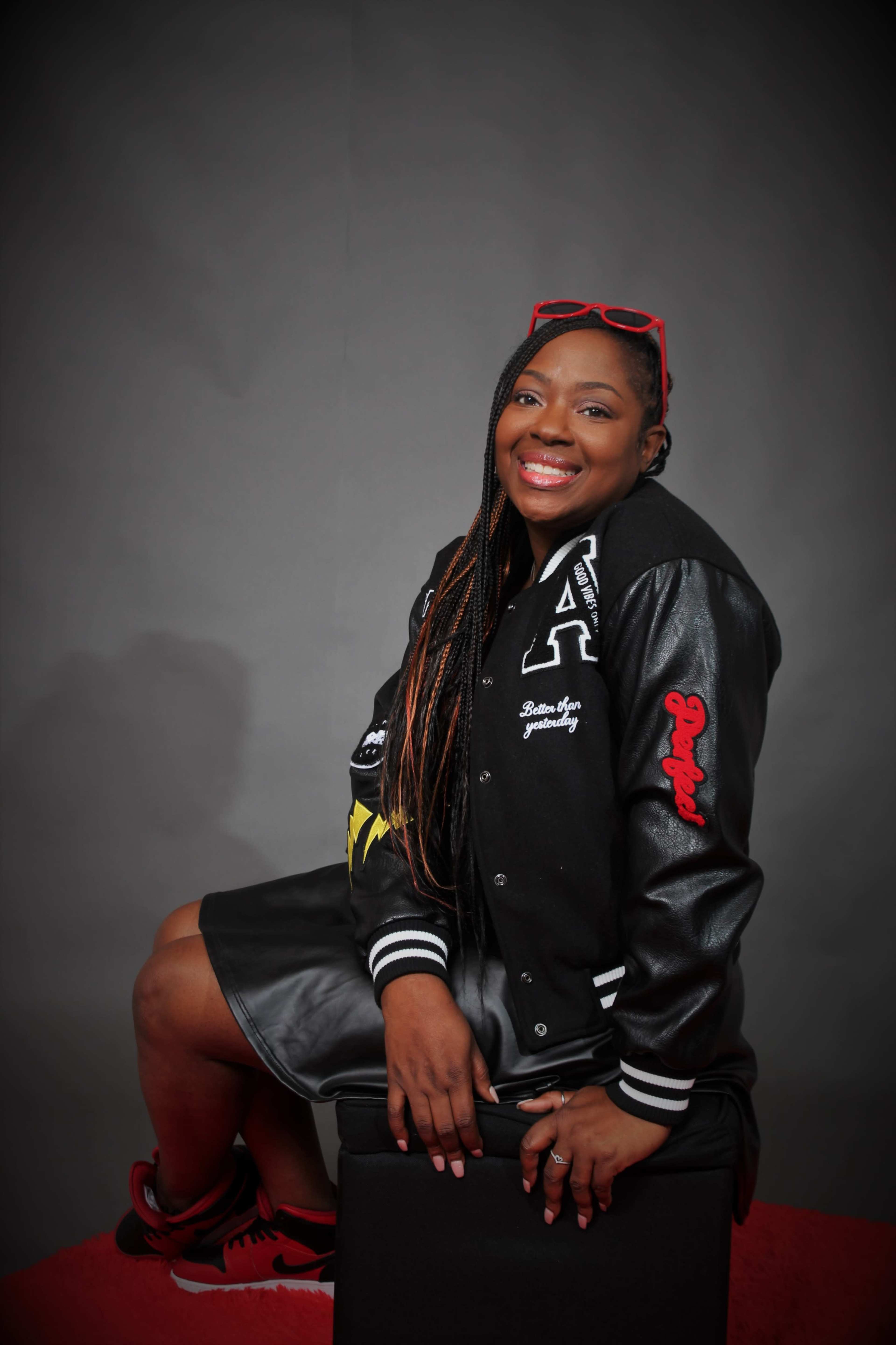 A woman with long braids, wearing a black varsity jacket and a leather skirt, sits on a black stool against a gray backdrop, smiling at the camera.