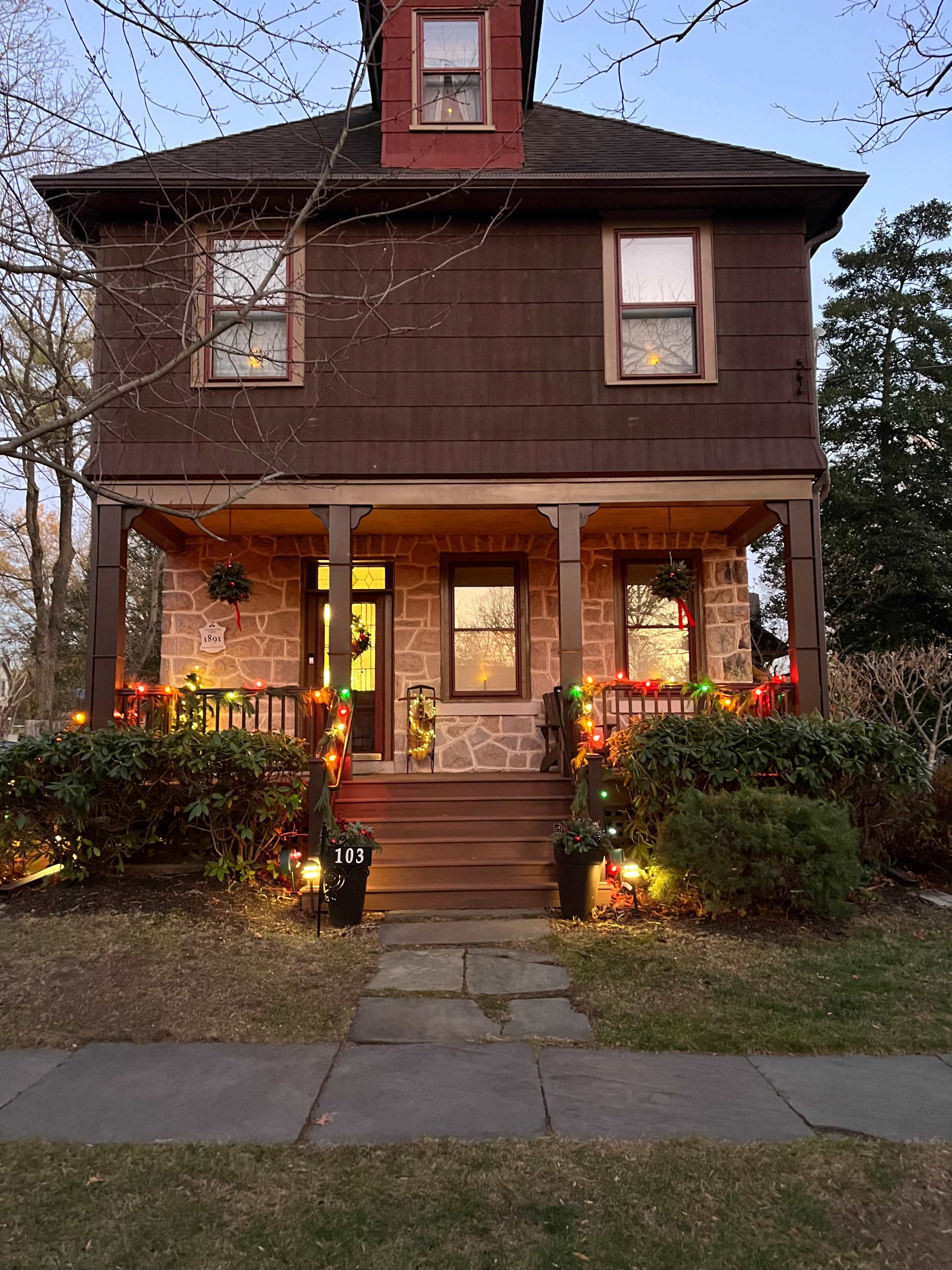 A two-story brown house with a red roof and wooden porch is decorated with multicolored lights for the holiday season.