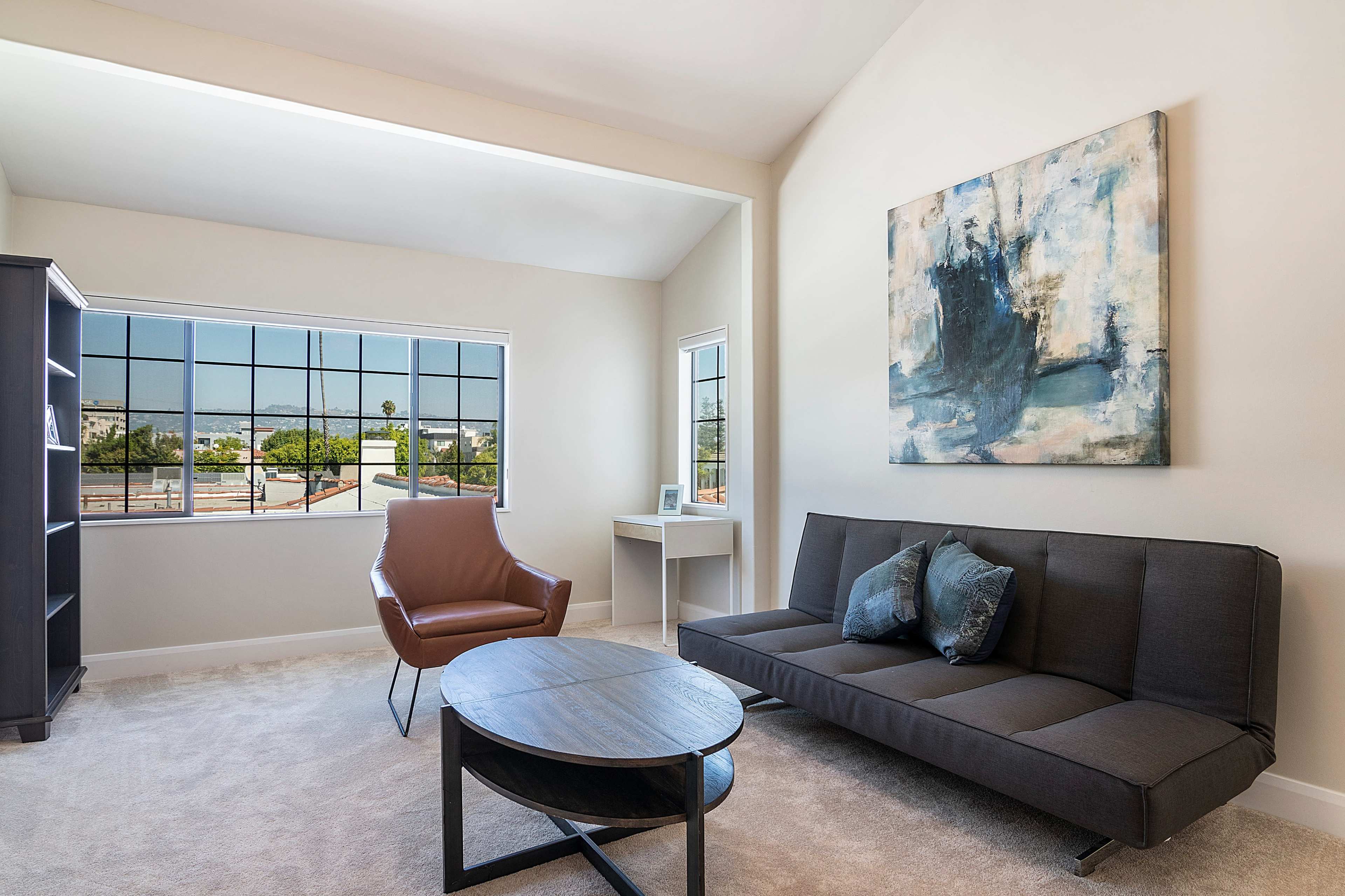 The image shows a modern living area featuring a black sofa, a brown armchair, a round coffee table, and a large window with a view of the outdoors.